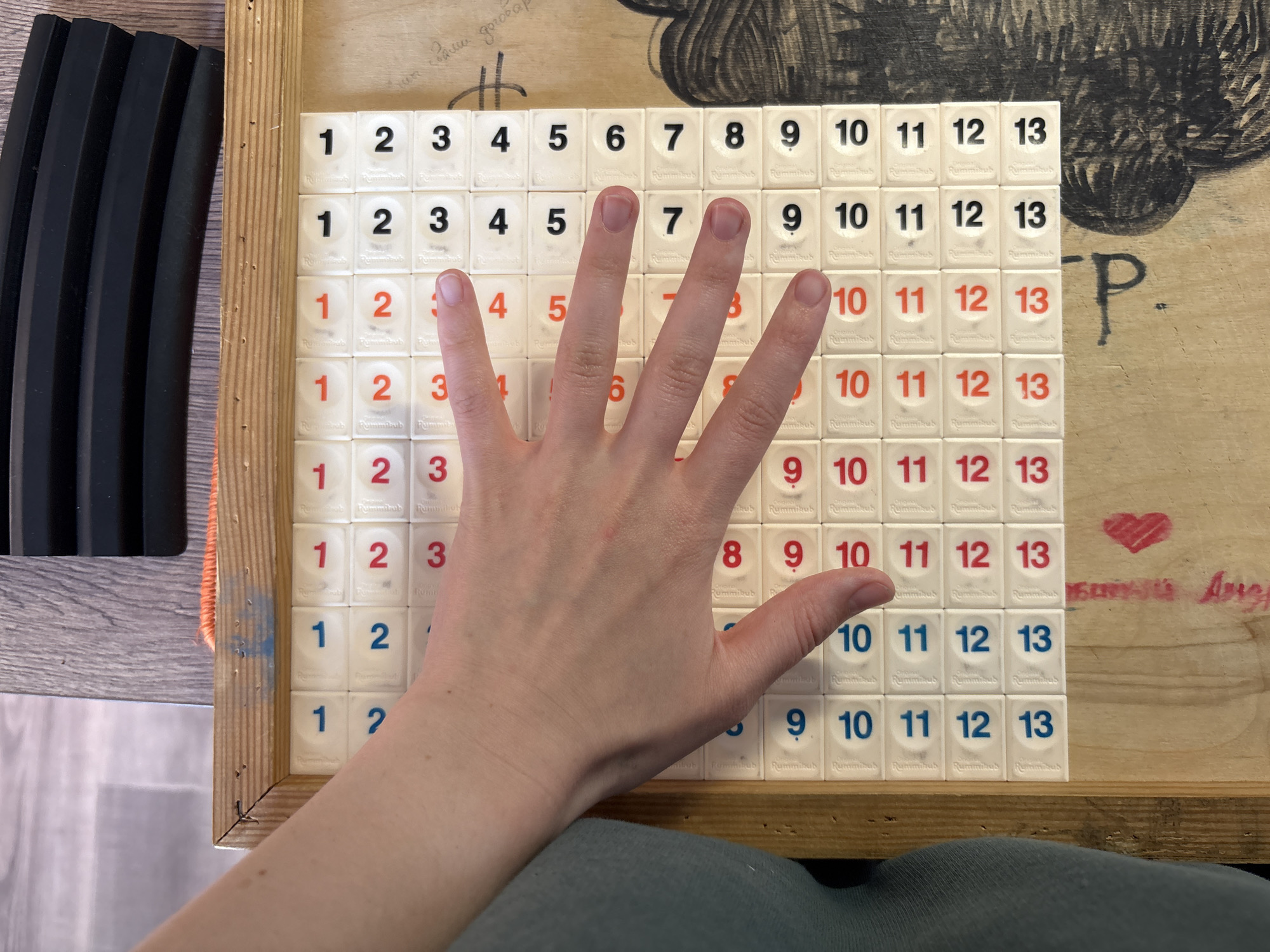 The image shows a person's hand placed over a Rummikub tile rack. The rack is filled with tiles numbered from 1 to 13 in both red and black. The hand is resting on the tiles, with the fingers spread out. The background includes a wooden table with some scribbled text and drawings. A black object with vertical lines is on the left side of the image. The person wearing gray pants is also visible.