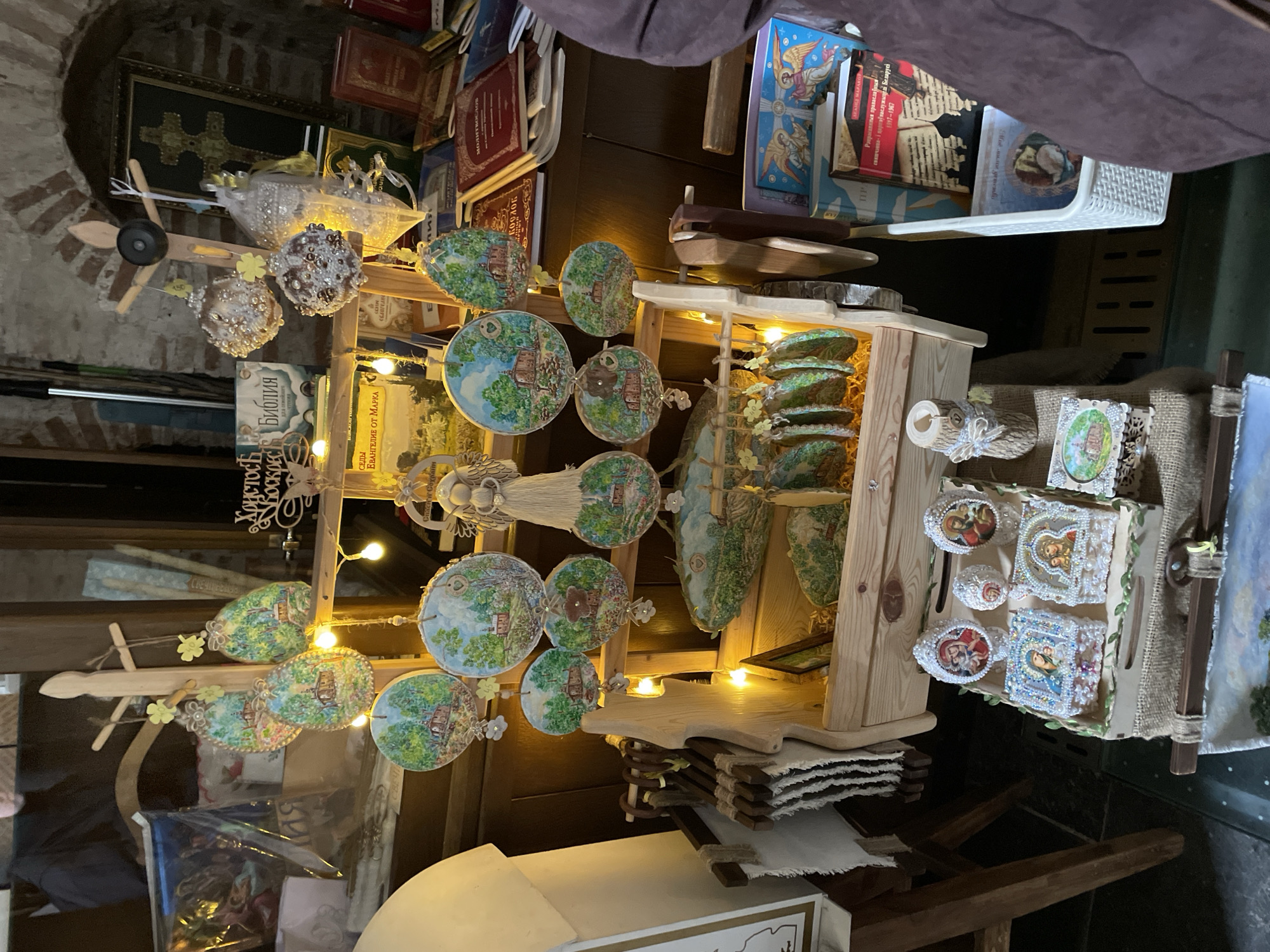 The image captures an indoor display of various crafted and religious items, likely in a gift shop, market, or religious store in Hrodna, Belarus. The scene features a central, multi-tiered wooden display stand illuminated by small string lights, creating a warm ambiance.
Hanging from the stand are numerous circular and egg-shaped ornaments, meticulously painted with idyllic green landscapes featuring houses and trees. Below these, a wooden model airplane hangs alongside two sparkly, spherical decorative pieces. On shelves within the main stand, there are stacks of small, intricately decorated boxes or miniature icons, some adorned with pearls or beads and depicting religious figures such as the Virgin Mary and the Christ child. Other sections of the display feature layered, green-colored decorative items, possibly carved wood or ceramic, resembling terraced landscapes, and small carved wooden logs with decorative flowers. The base of the display holds stacks of thin wooden boards and rolled-up fabric pieces.
The background reveals shelves packed with books, many bearing titles in Cyrillic script. A framed image of a cross is visible against a brick wall in the upper left, suggesting an older building. Visible text on the books includes "БИБЛИЯ" (Bible), "ЕВАНГЕЛИЕ ОТ МАРКА" (Gospel of Mark), "Спаси и Сохрани" (Save and Protect), and "Христосъ Воскрес" (Christ is Risen), all pointing to a strong Eastern Orthodox religious context. One book title reads "Рапраганяныя праваслаўны імпелі Беларус 1917-1967" (Dispersed Orthodox Impulses of Belarus 1917-1967), further connecting the display to Belarusian Orthodox culture and history. The original image is rotated 90 degrees counter-clockwise.