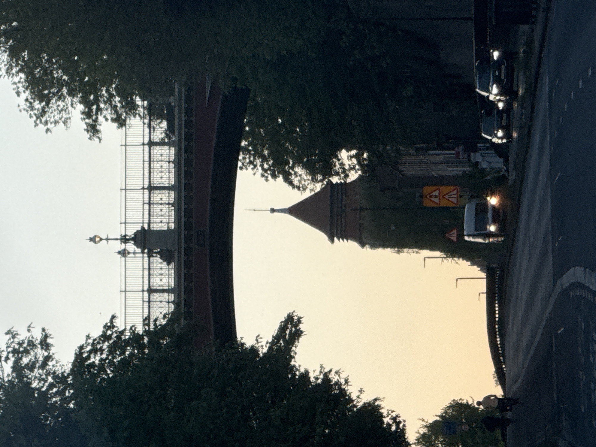 The image, oriented correctly, depicts an urban landscape, likely in London, United Kingdom, during either sunrise or sunset. The scene is dominated by a large, ornate bridge with intricate metal railings and decorative lamp posts on the left, and a distinctive tower with a conical roof and spire in the middle-ground.
On the right side of the frame, a paved road carries vehicles, including at least two dark cars and a white van, all with illuminated headlights, indicating low light conditions. A single person is visible cycling along a path adjacent to the road, appearing to be shirtless or in light-colored clothing.
The sky is a warm, diffused light, transitioning from a yellowish-orange glow near the horizon to a lighter, softer tone higher up, silhouetting most of the foreground elements such as dense trees. A yellow diamond-shaped road sign with two black exclamation marks (a general warning symbol) is visible next to the road, behind the white van. The overall atmosphere suggests an early morning or late evening commuter scene.