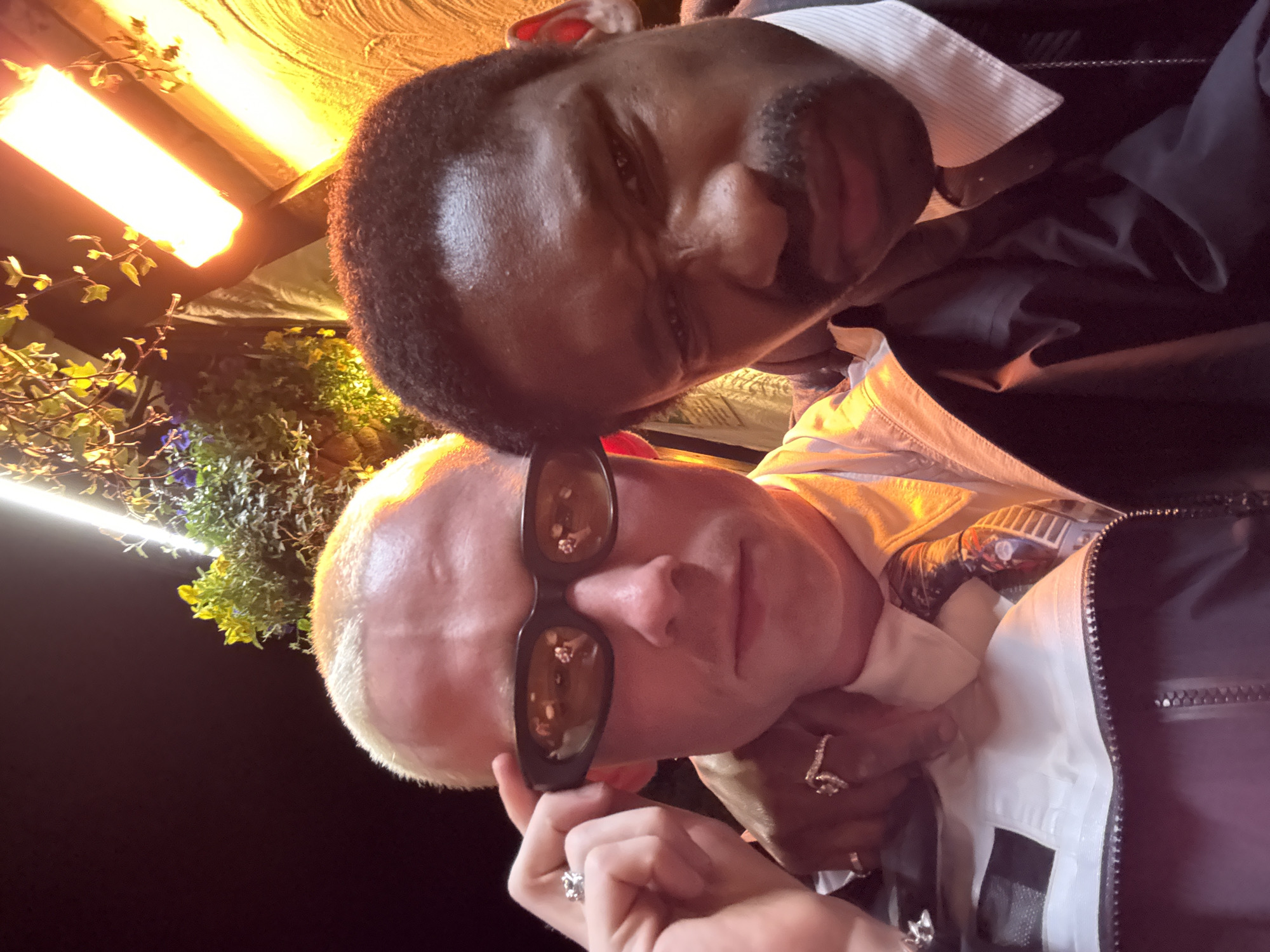 The image captures two individuals in close proximity, likely a selfie or a photo taken at close range. The person in the foreground has fair skin and light-colored hair, wearing sunglasses. Their hand is visible, adorned with a ring. The other person has dark skin and short, dark hair with a mustache. They are wearing a light-colored collared shirt beneath a dark jacket. The background suggests an outdoor setting, with greenery and some artificial lighting. This image was likely taken in London, United Kingdom.