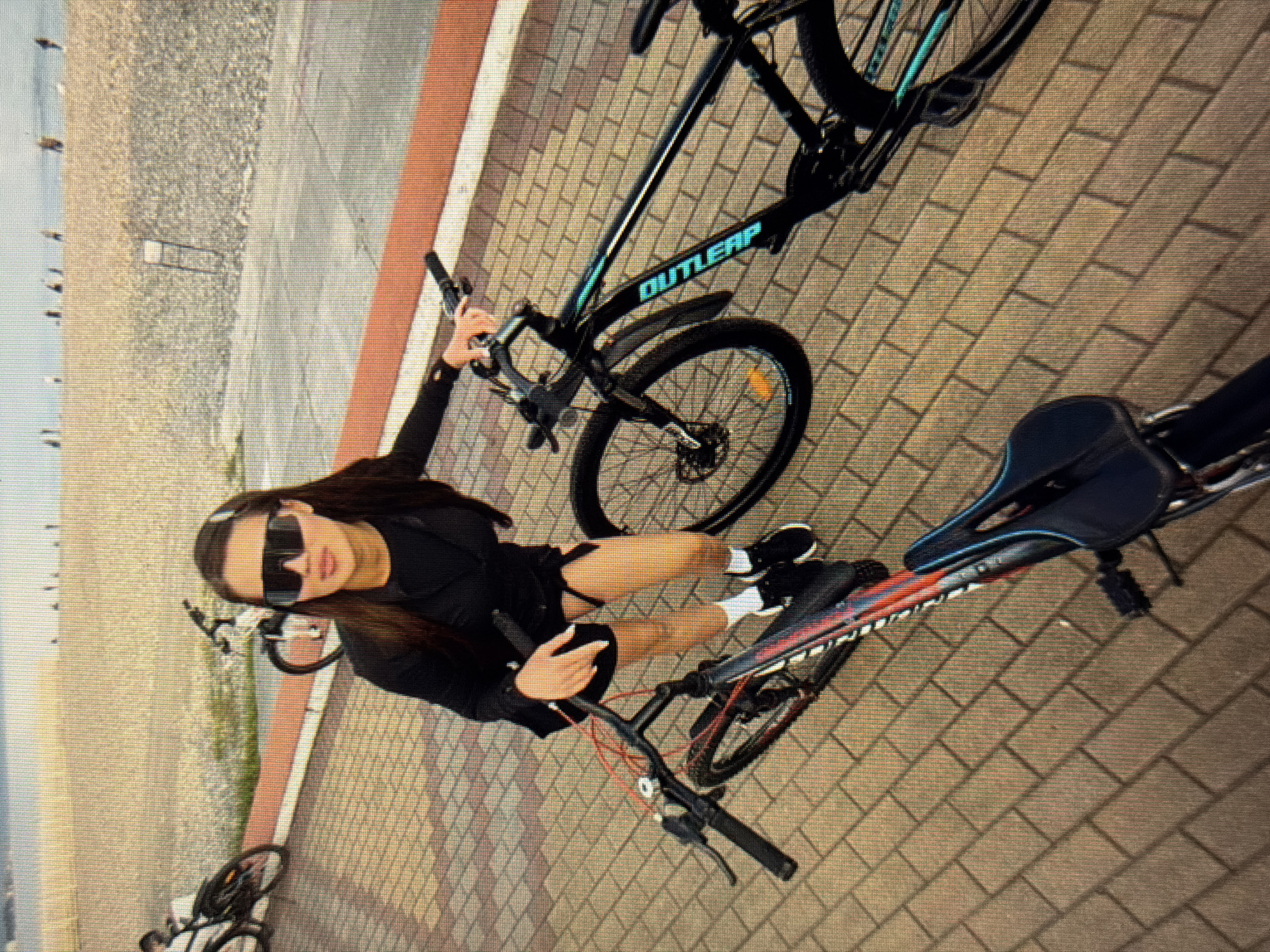A woman wearing sunglasses and a black jacket over a black outfit stands with two bicycles on a brick-paved promenade. One bicycle, black with teal lettering, is to her right. The other bicycle, with a red and black frame, is positioned in front of her. In the background, a beach with scattered people and the sea can be seen. The location is identified as Sirius, Russia.