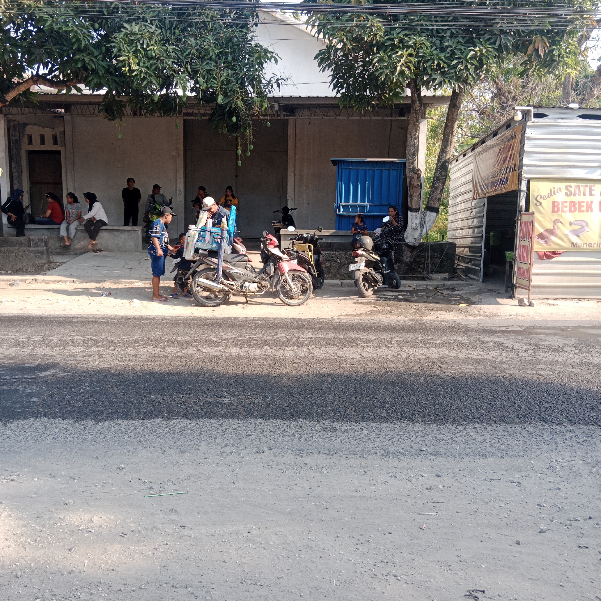 A group of people are gathered near a row of buildings and parked motorcycles in Tegalsari, Indonesia. The buildings appear to be small shops or eateries, with one prominently displaying a sign advertising "Sate Bebek" (duck satay). Several individuals are seated on a low wall in front of one building, while others stand or lean against motorcycles. Two children are interacting in the foreground, with one appearing to be serving something from a cart attached to a motorcycle. The environment is a street scene with a paved road and a dirt shoulder. Trees with visible fruit hang over the buildings. The time of day appears to be daytime, with sunlight casting shadows.