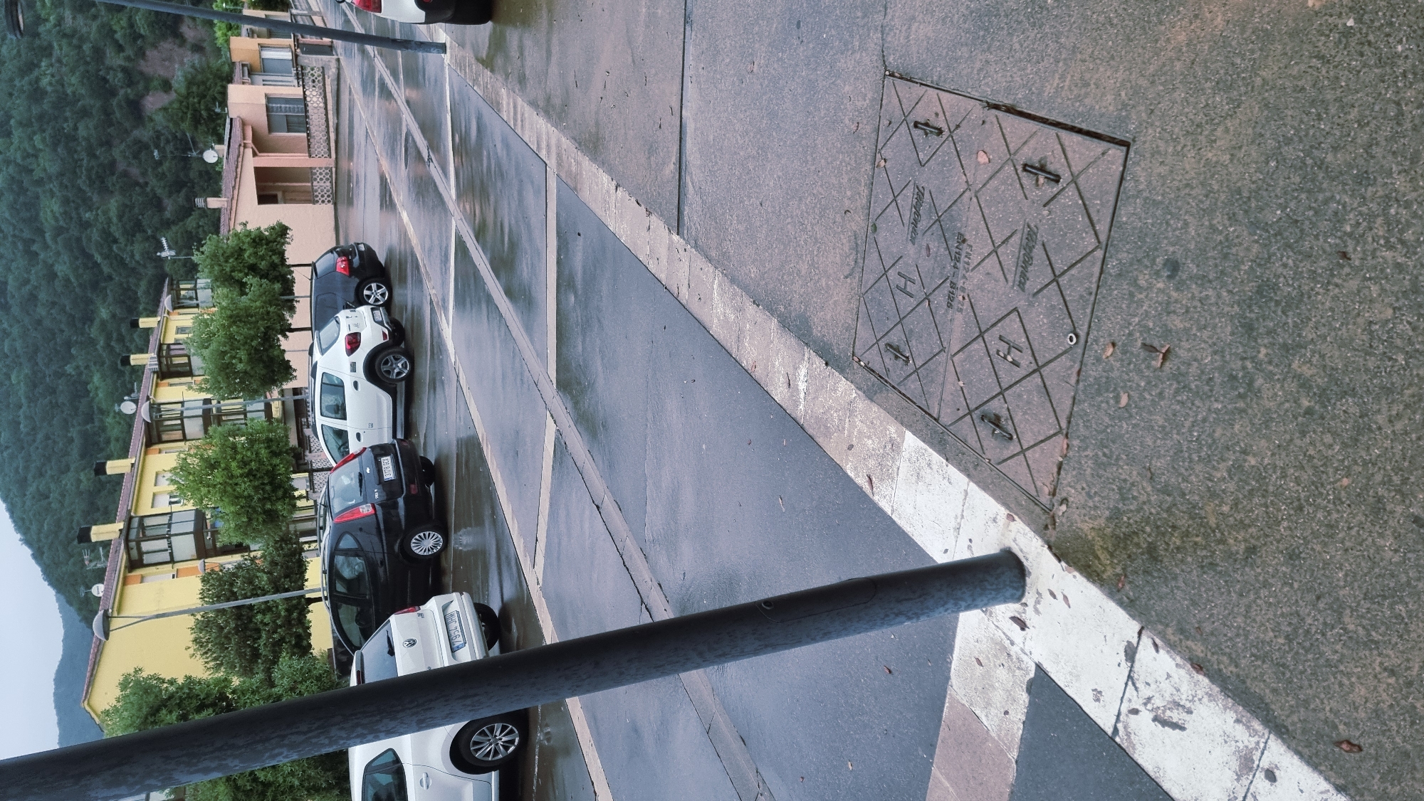 The image depicts a street scene in Sant Joan Les Fonts, Spain, on a wet, overcast day. The foreground is dominated by a manhole cover with a diamond pattern and markings that include "H" and "Telifon," suggesting it is part of a telecommunications infrastructure. Adjacent to the manhole cover is a grey concrete sidewalk. A large, dark metal pole, likely a street lamp, stands prominently on the left, partially obscuring a white-lined parking space. Beyond the pole, several cars are parked on a wet asphalt surface. These include a white Volkswagen Polo on the left, a black minivan with license plate "3179 GCJ" in the center, a white SUV, and a black car on the right. In the background, a row of yellow apartment buildings with balconies lines the street, backed by a densely forested hillside. The overall impression is a quiet, everyday urban setting experiencing inclement weather.