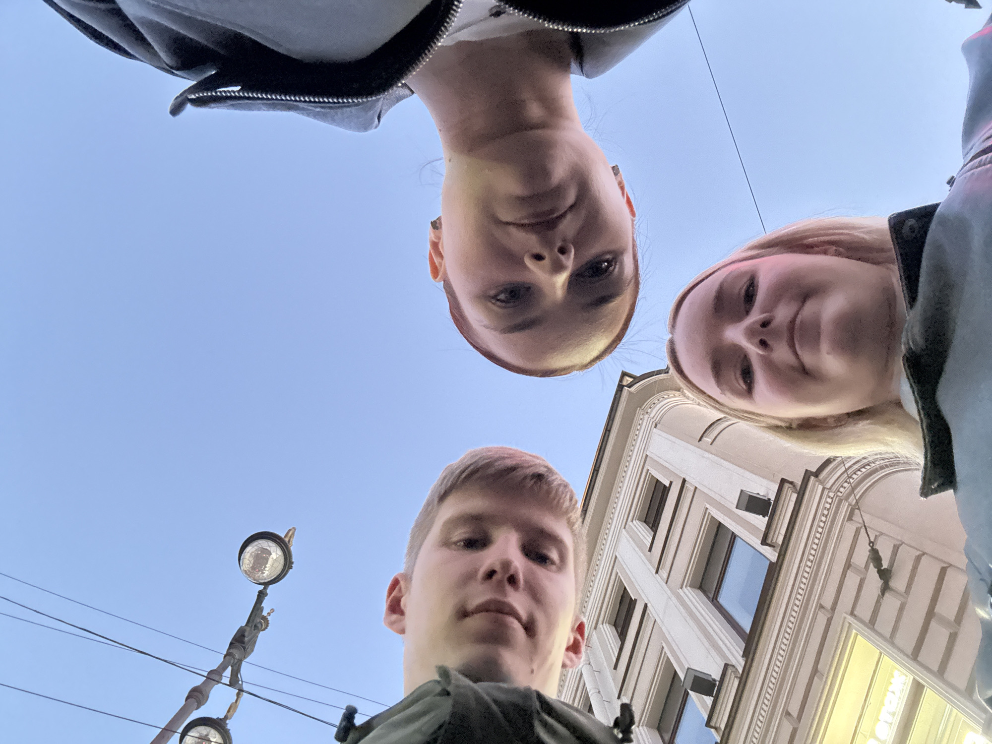 A low-angle selfie captures three individuals looking down at the camera against a clear blue sky. A building with decorative molding and windows, characteristic of Saint Petersburg architecture, is visible on the left. A street lamp and electrical wires are also present in the upper portion of the frame. The people are positioned with their heads close together, suggesting a shared moment or activity. One individual on the left has fair skin and light hair, looking directly at the camera with a slight smile. Another individual in the center has fair skin and reddish-brown hair, also looking down. The third individual on the right has fair skin and light blonde hair, smiling at the camera. All three appear to be young adults. The overall atmosphere is casual and spontaneous.