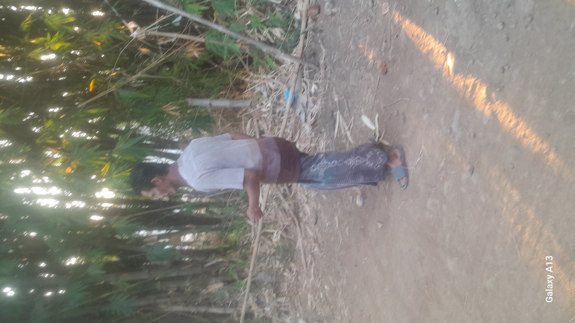 A man stands in a dirt path, partially obscured by trees. He is wearing a light-colored short-sleeved shirt with a darker horizontal band around the waist, and a patterned sarong. He is facing away from the camera, looking to his left. He holds a thin stick or pole in his right hand. The ground is covered in dirt and scattered dry leaves and twigs. In the background, a dense stand of bamboo trees is visible, with sunlight filtering through the leaves. The overall scene appears to be in a rural or semi-rural outdoor environment. The timestamp "Galaxy A13" is visible in the lower-left corner.