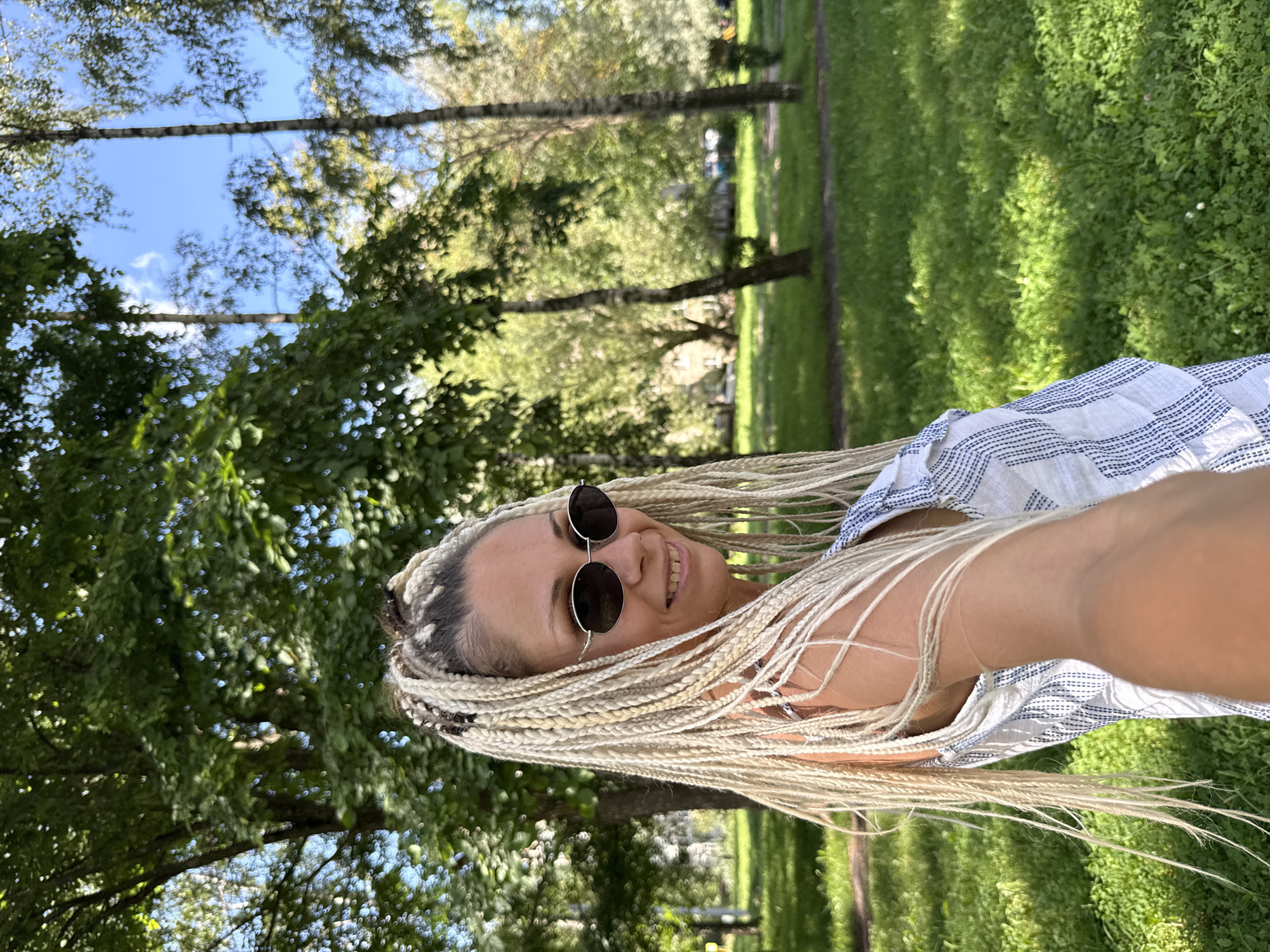 A woman with blonde box braids and sunglasses stands in a park in Rusanovo, Russia. She is wearing a white top with blue stripes. The background is filled with green trees and grass, and the sky is blue with some clouds. The woman is smiling and looking at the camera. The photo appears to be a selfie.