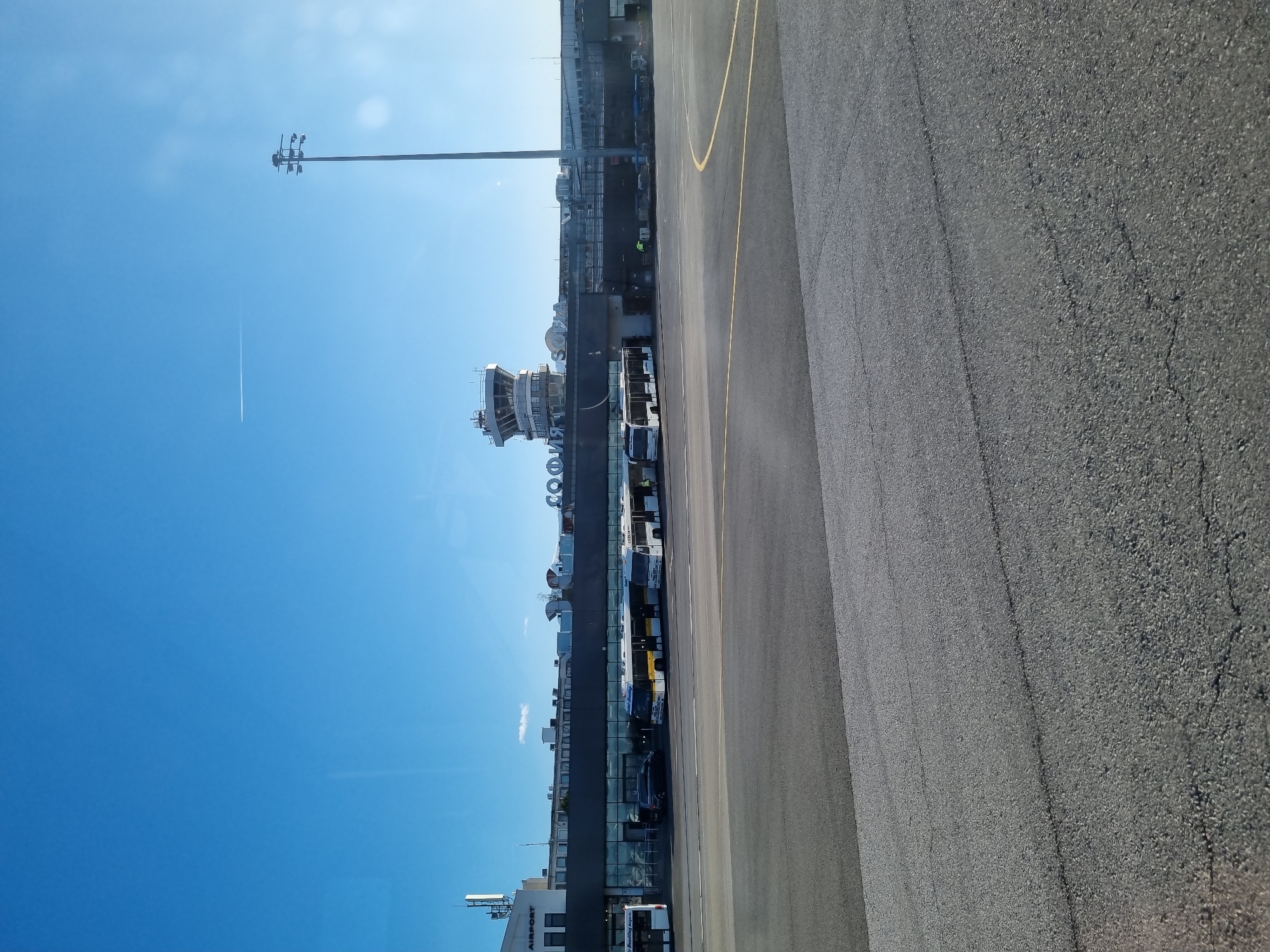 The image depicts a section of Sofia Airport in Bulgaria. In the background is the airport control tower, standing adjacent to the airport terminal building. Adjacent to the terminal, multiple shuttle buses and vehicles are parked, presumably transporting passengers between the terminal and aircraft. In the foreground, there's a stretch of asphalt, marked with a yellow line, likely part of the taxiway or apron. The sky is blue with a clear contrail from a plane.