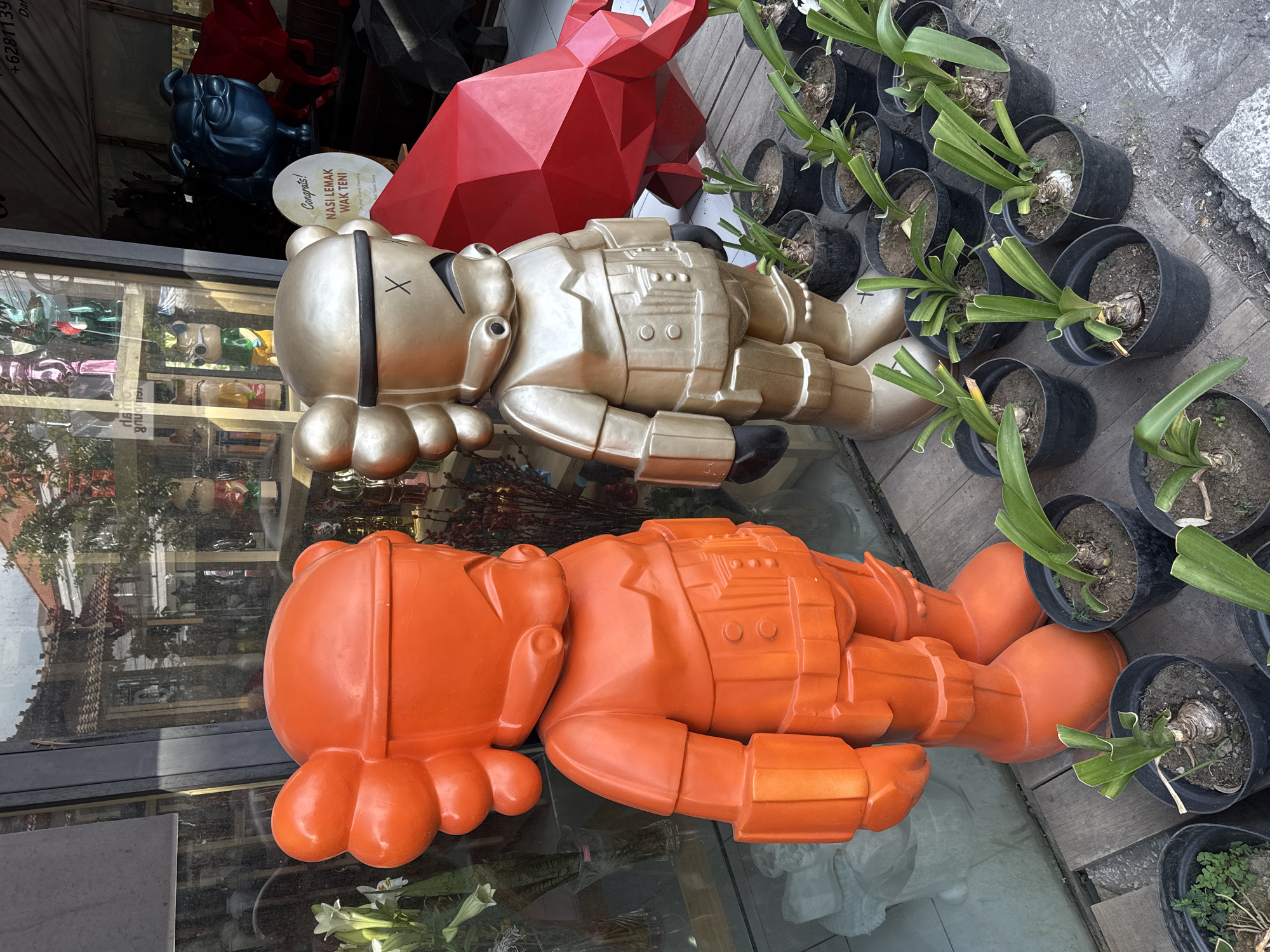 Two large, stylized figures, one bright orange and the other metallic gold, stand prominently on a wooden platform outside a building. The figures resemble cartoon characters with large, bulbous heads and detailed, armor-like bodies. The orange figure stands on the left, while the gold figure is to its right. In the background, visible through large windows, are shelves filled with other colorful figurines and decorative items. To the right of the figures, a large, faceted red sculpture resembling a bull is partially visible. In front of the figures and scattered across the platform are several black potted plants with green, strap-like leaves. The scene appears to be a retail storefront, possibly a gallery or boutique, located in North Kuta, Indonesia. The environment suggests an outdoor display area in a commercial district, with a building facade and reflections in the glass.
