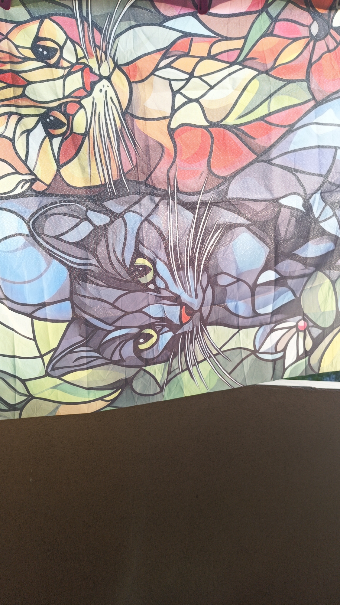 The image shows a colorful, patterned fabric or print featuring two cat illustrations in a stained glass style. One cat is orange and yellow, and the other cat is black and blue. The lower portion of the image is a dark brown, possibly a solid surface that the fabric is placed on. There is no indication of the location being Karnobat, Bulgaria, or any other specific location.