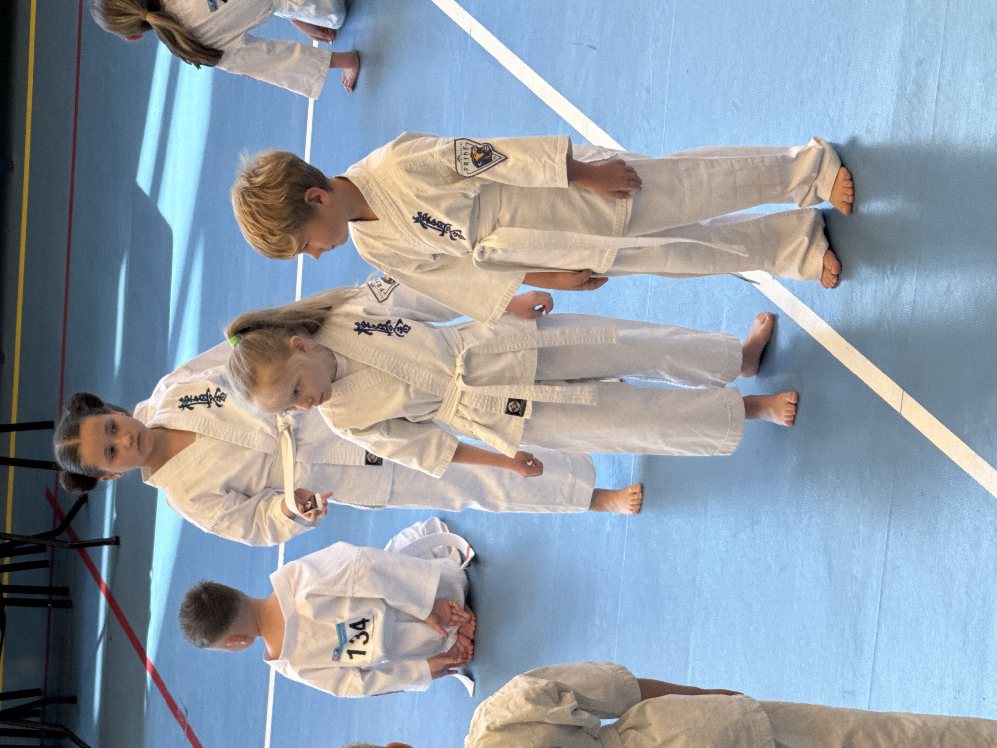 Several children wearing white karate uniforms stand and kneel on a blue floor with white, red, and yellow lines, potentially a gymnasium or training hall. One child kneels and has the number "134" displayed on their back. The children are barefoot. The uniforms have patches on the sleeves with the word "PRIDE" on them. Some children are interacting with their belts. The location is unknown.
