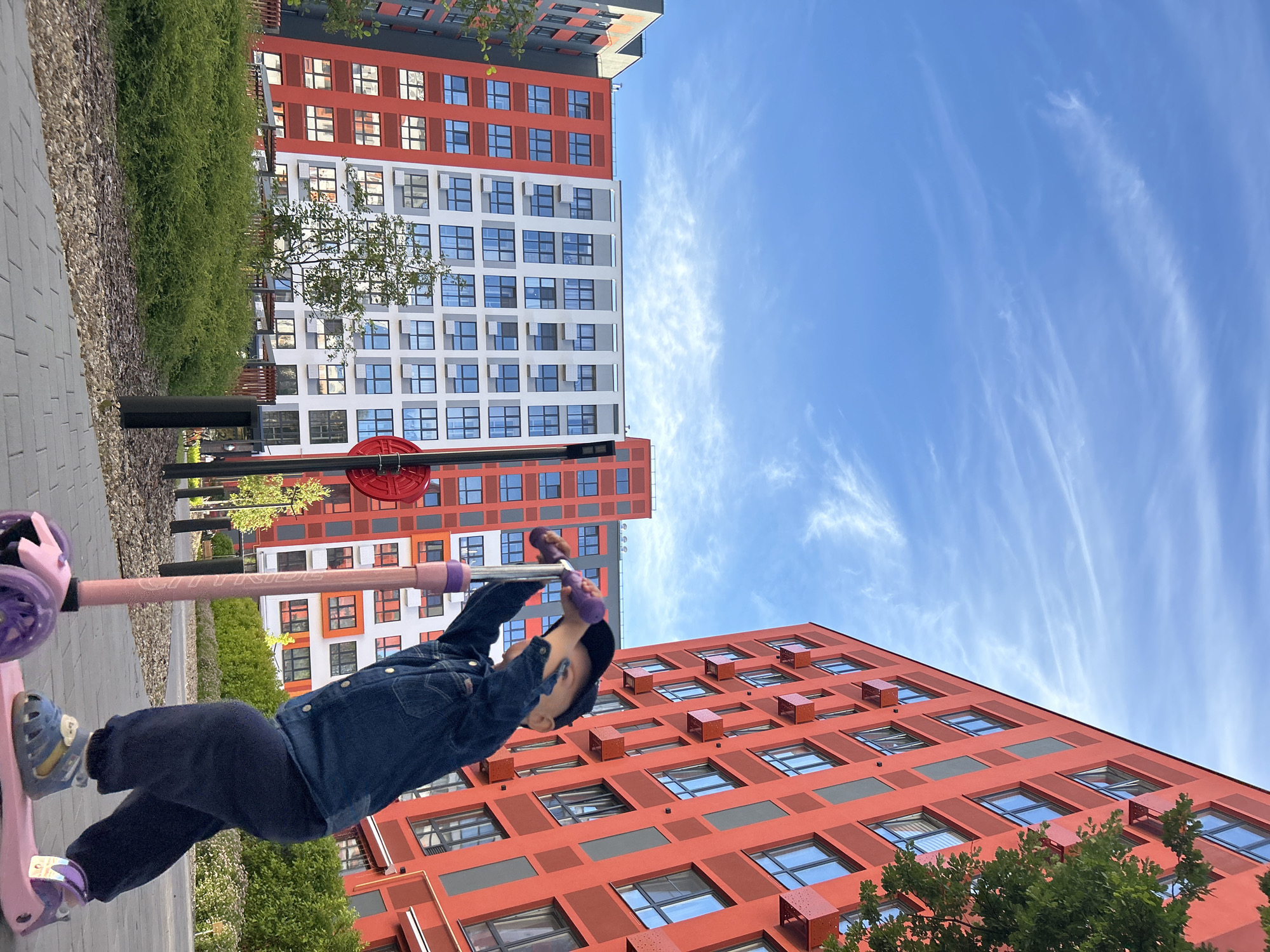A young child rides a pink scooter with purple accents on a paved path. The child is wearing a denim jacket, dark pants, and blue sandals. In the background, there are two modern apartment buildings. One building is primarily white with red accents, and the other is predominantly red with dark gray accents. Both buildings have numerous windows. The sky is bright blue with wispy white clouds. The scene suggests a sunny day, likely late afternoon or early evening given the angle of the light. The setting appears to be a residential area or courtyard in Kaluga, Russia, with greenery visible between the buildings. The child's posture and the overall composition imply an active and playful moment. The word "GRIT" is visible on the scooter's handlebar.