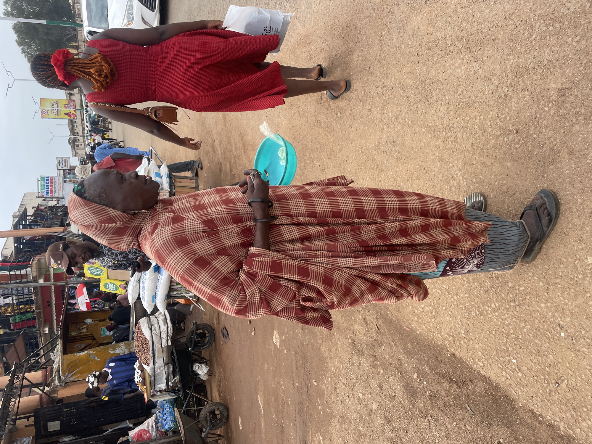 A man in a red plaid robe and sandals stands on a dirt road in Jos, Nigeria. He holds a small blue bowl in his left hand and a cigarette in his right hand. In the background, a woman in a red dress walks away from the viewer. Street vendors are present along the road, selling goods from carts. Buildings with signs, including one for "TRUSTCARE MEDICAL LABORATORIES", line the street. A white car is parked in the background.