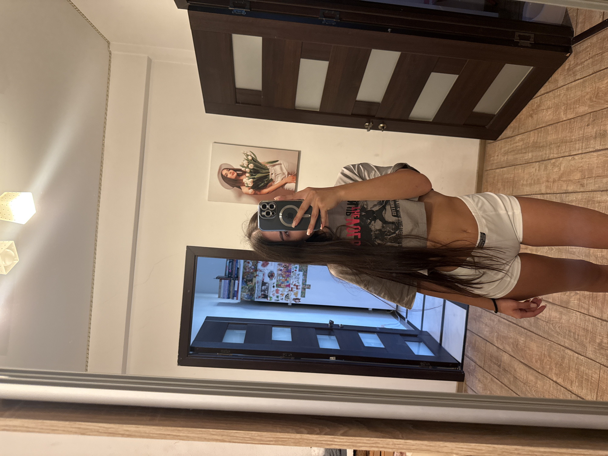 The image shows a young woman taking a mirror selfie in an indoor environment, likely a residential room. She is wearing a grey cropped t-shirt and white shorts. Her long dark hair covers part of her face as she holds a smartphone to capture the reflection. In the background, there are two wooden doors, one partially open revealing a glimpse of a room with shelves filled with books and decorative items. On the wall between the doors, a framed picture of a woman holding tulips is visible. The floor is made of wooden planks. The overall scene suggests a casual setting within a home. There is no information in the image to suggest it is located in Chișinău, Moldova.