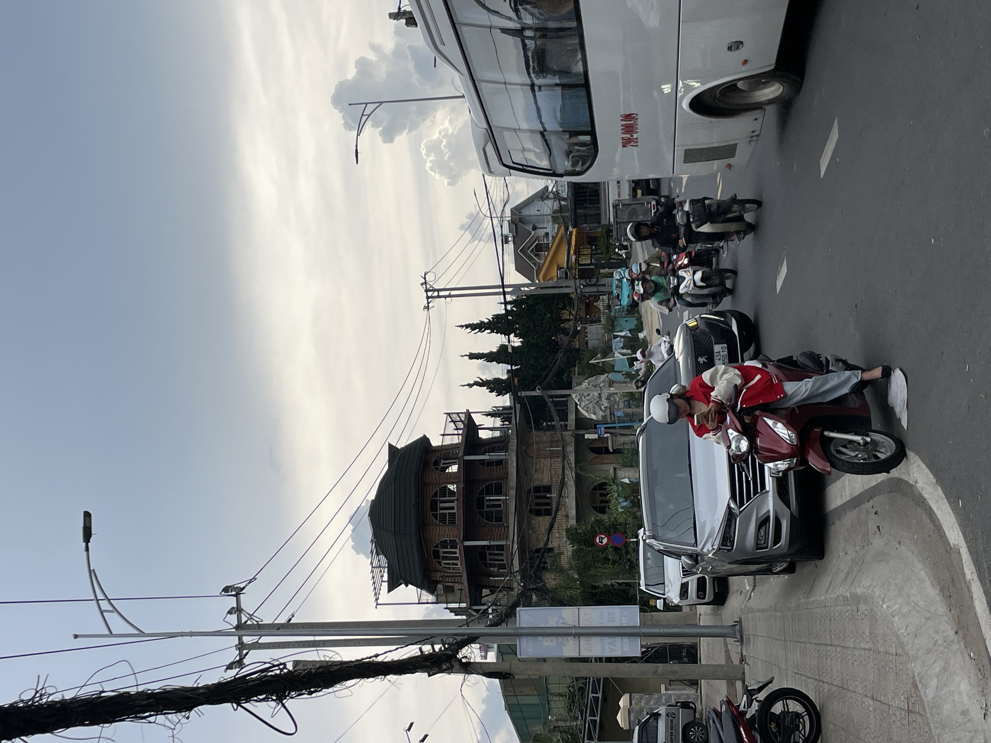 The image shows a street scene in Lâm Viên - Đà Lạt, Vietnam, characterized by a unique, multi-tiered building with a distinctive curved, dark green roof. The building appears to be made of stone and wood, with arched windows and balconies. In the foreground, a woman on a red scooter is looking at her phone, wearing a white helmet and a red and white jacket. She is stopped at the side of the road, next to a silver SUV. Several other motorbikes and cars are on the street, suggesting moderate traffic. A large white bus is also visible, with red lettering on its side. Overhead, power lines crisscross the sky, and a streetlamp stands tall. The sky is partly cloudy, with dramatic cloud formations in the background. The overall impression is one of a busy urban environment with architectural and natural elements.