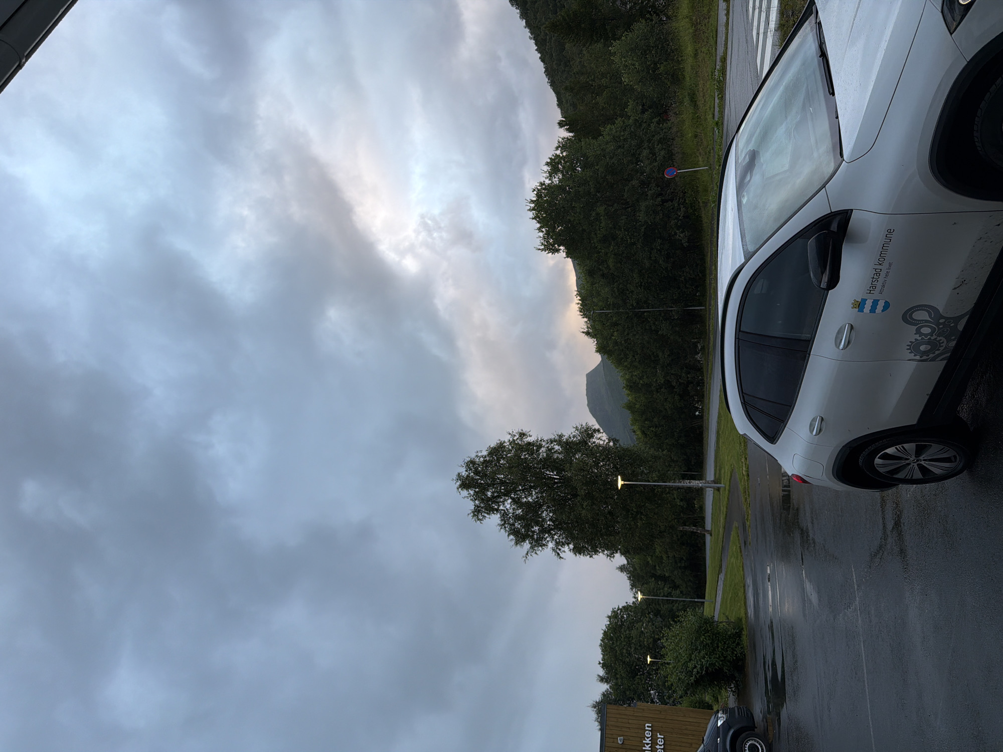 A white Mercedes-Benz GLA SUV with the Harstad kommune logo and text "Attraktiv hele livet" is parked on a wet asphalt lot. The lot is bordered by a grassy area with trees and streetlights, and further back by a densely wooded hillside. A dark gray van is visible on the left side of the frame. In the background, a mountain peak is visible through the trees under a cloudy, overcast sky. The scene appears to be in the city of Harstad, Norway, likely a municipal parking area.