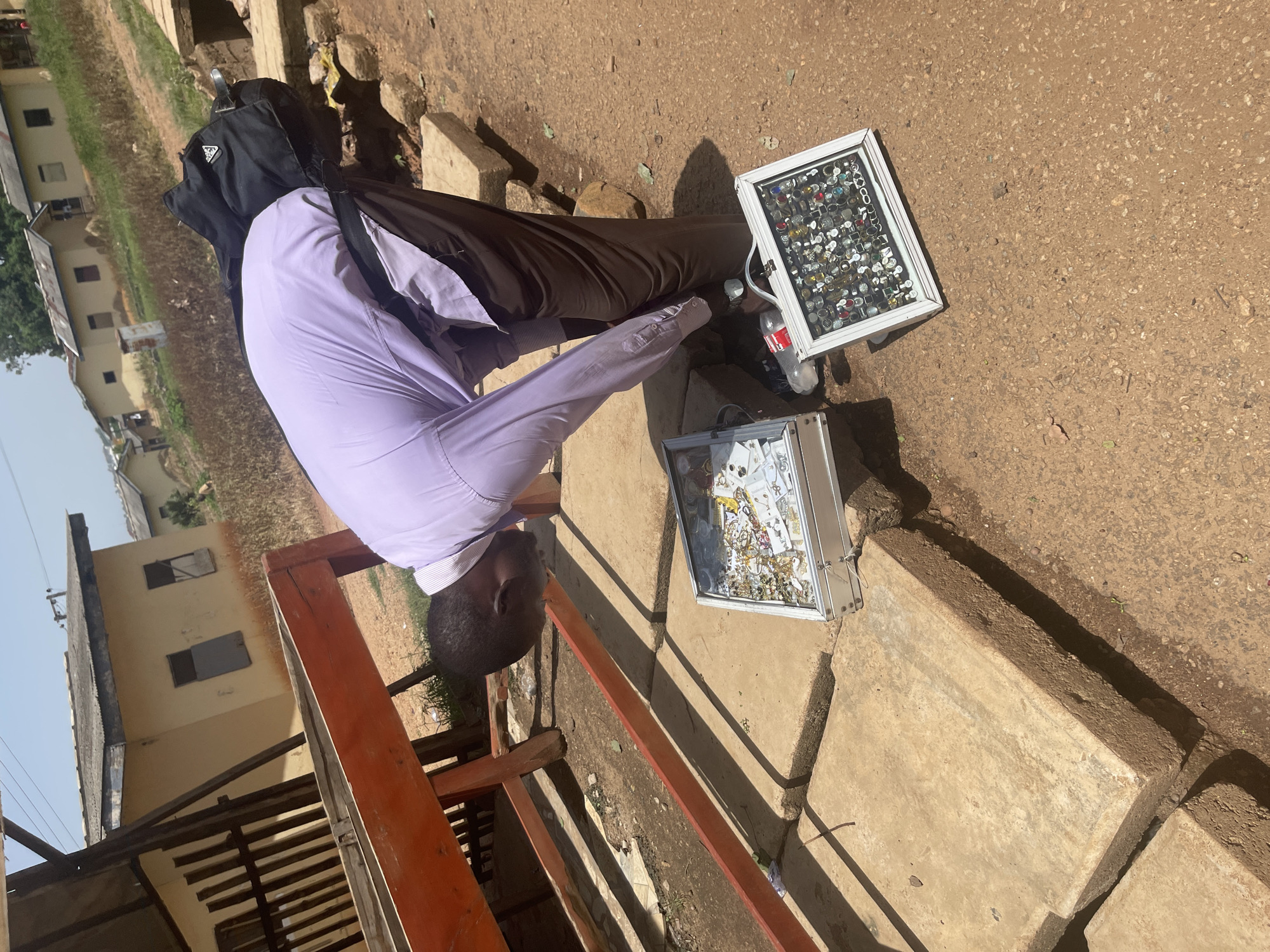 A man, wearing a lavender dress shirt and brown trousers, is bent over two display cases of jewelry placed on concrete blocks in Rafin Par, Nigeria. The display cases are made of silver-colored metal. One case, closer to the man, contains necklaces and bracelets. The other case, further away and at an angle, is filled with rings of various designs. A black backpack is slung over the man's shoulders. The background shows a dirt path, vegetation, and several small buildings with corrugated metal roofs, suggesting a modest urban or semi-urban setting. The sky is clear and bright.