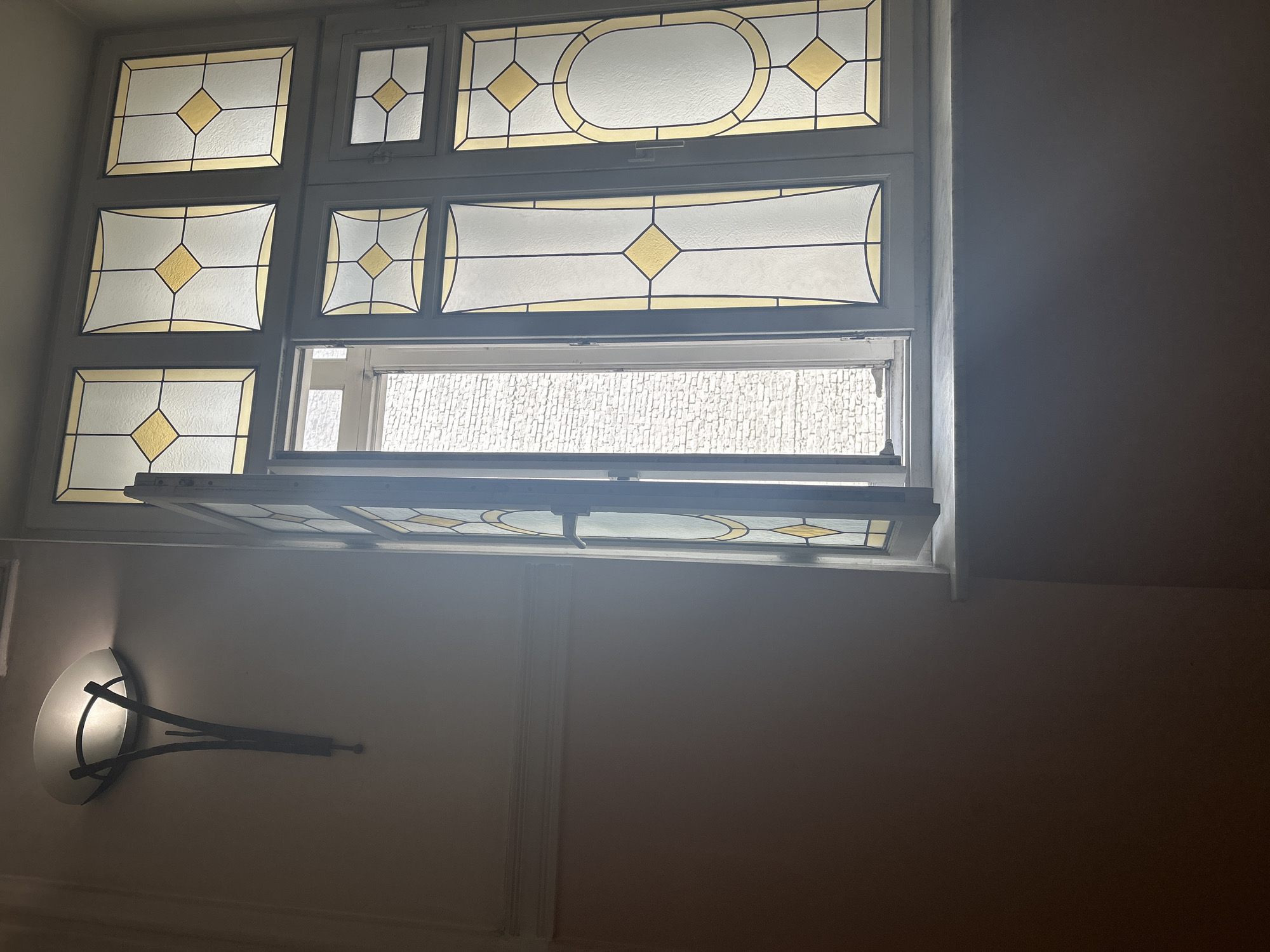 The image shows a window with decorative stained glass in a room with neutral-colored walls. The window is partially open, revealing a view of an exterior wall with a textured surface. Below the window, on the wall, is a sconce-style light fixture. The overall lighting in the room is dim, with the right side of the image appearing darker.
