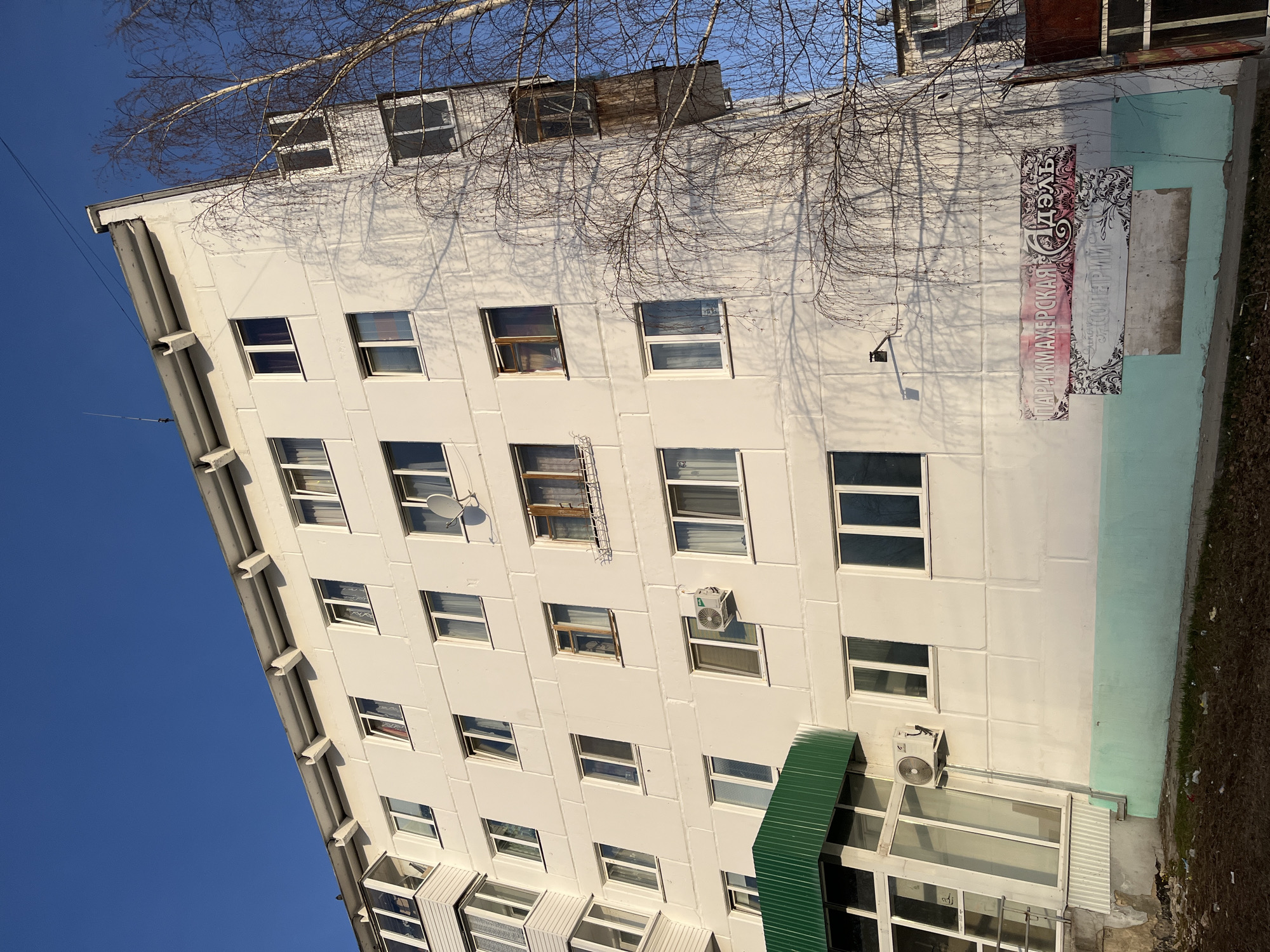The image shows a multi-story, white apartment building, likely located in a city or town based on the presence of urban infrastructure. The building has a grid-like facade due to its repeating rectangular windows. There are some satellite dishes mounted on the exterior wall. The building is adorned with an array of windows. Additionally, what appears to be a sign or advertisement is affixed to the side of the building. A tree is located nearby, with its bare branches visible in the foreground and casting shadows on the building's facade. The sky is a clear, bright blue, indicating a sunny day. Based on information available, the location is likely in the city of Chaikovsky, Russia.