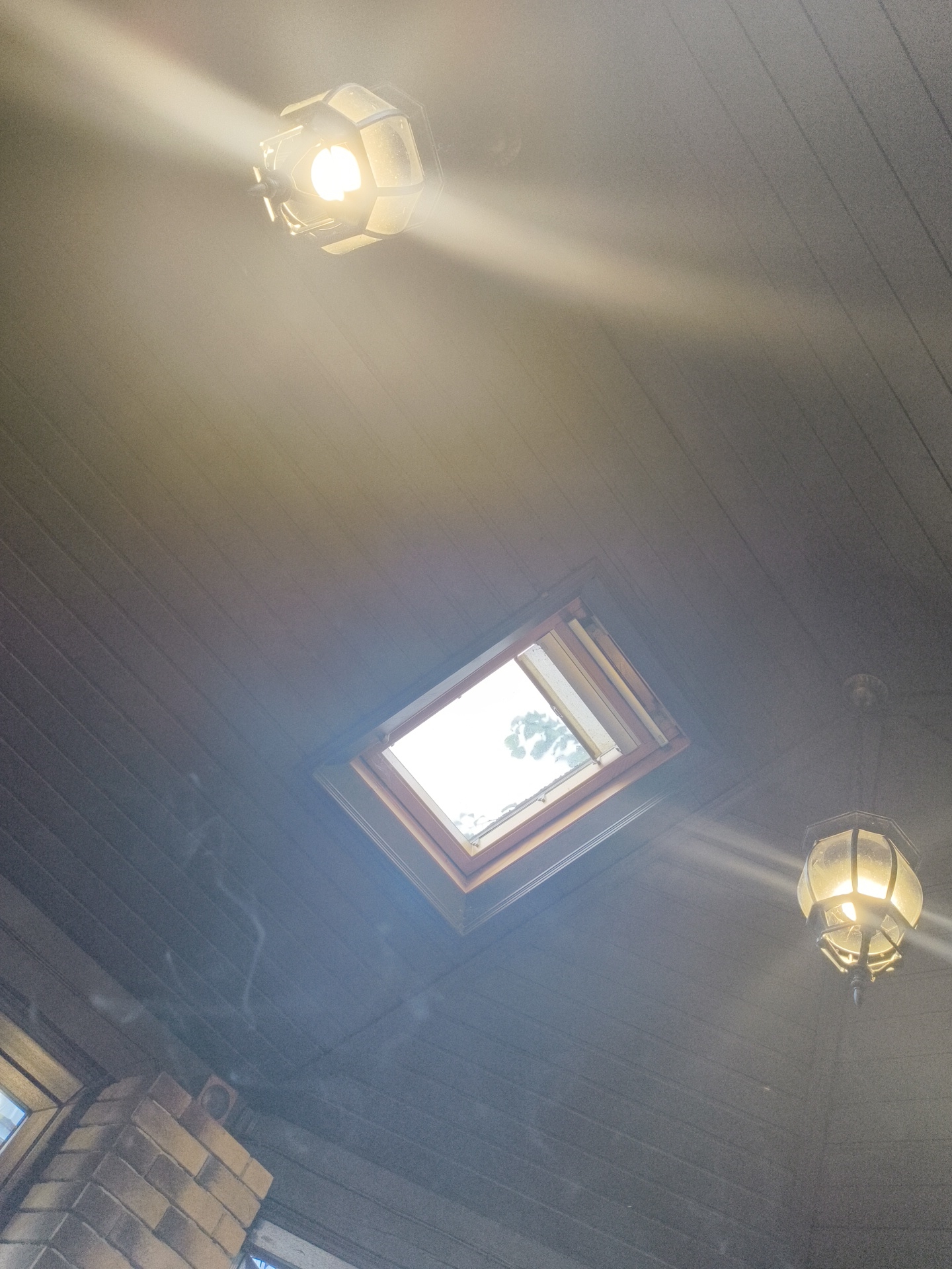 The image shows an interior ceiling with a wooden plank design. There is a square skylight in the center of the ceiling, letting in natural light. Two pendant lights are also visible, one on the upper left and another on the lower right of the image. Both lights are illuminated and cast strong beams of light. There is a brick structure, possibly a chimney or part of a wall, in the lower left corner of the image. Some faint wisps of smoke are visible in the air, particularly in the left and center portions of the image. The overall lighting is bright due to the skylight and the lamps, creating some lens flare and haze. The location is identified as Востец Снт, Russia.