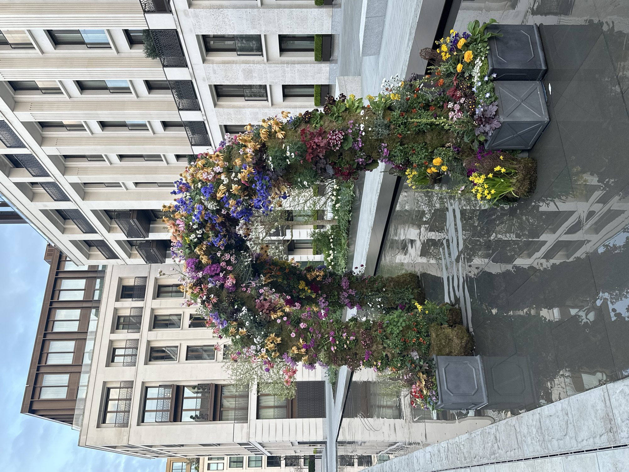 The image shows a floral archway or installation composed of various flowers and greenery, with a reflection in the water-covered pavement. The installation is set against the backdrop of modern buildings with light-colored facades and dark window frames. The buildings are several stories high and feature balconies with decorative railings. The sky is partly cloudy. The installation appears to be located in a public space, possibly a courtyard or square in a city setting. Given the architectural style of the buildings, this could be located in London, United Kingdom.