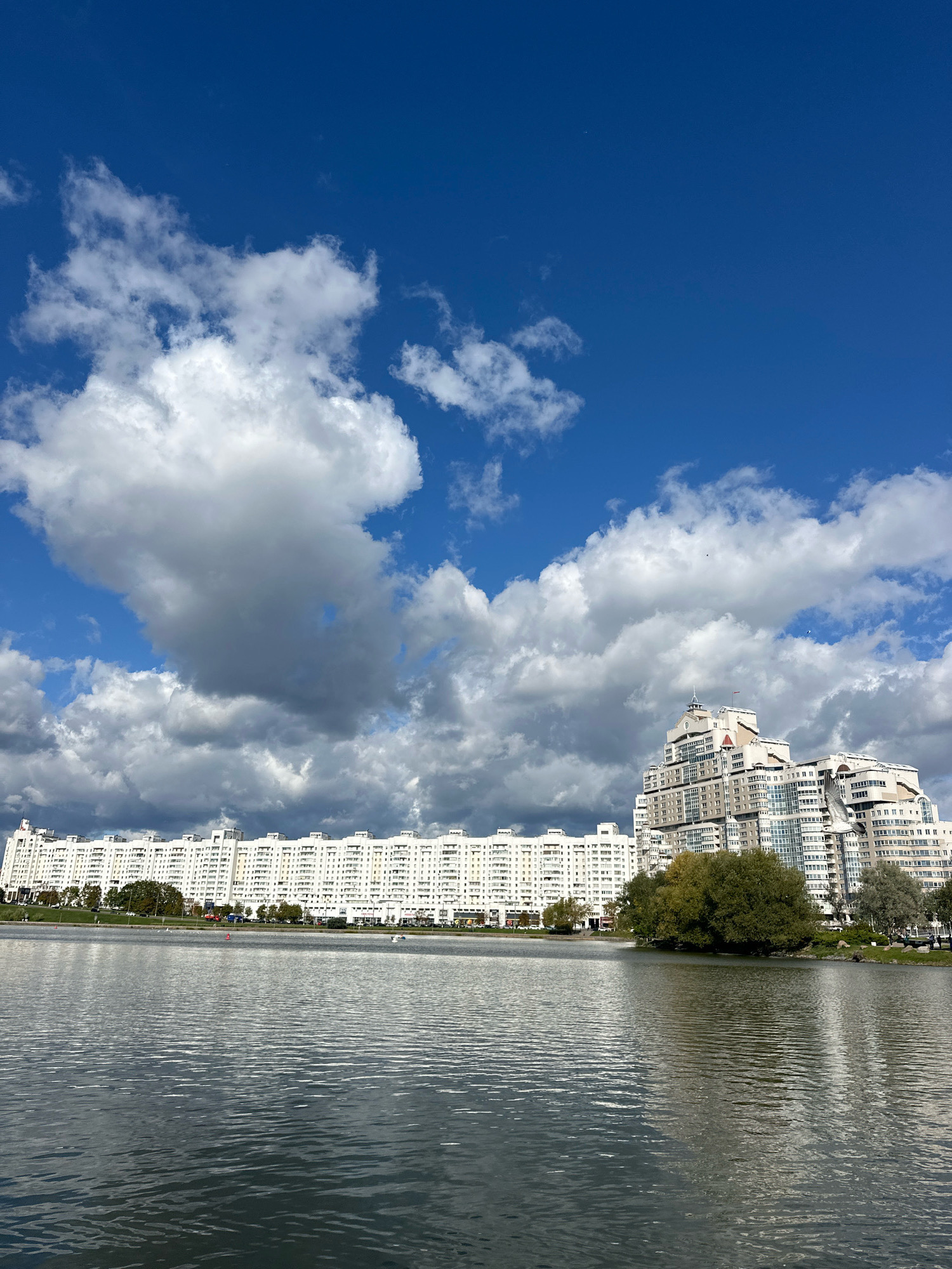 The image captures a wide view of the Svislach River in Minsk, Belarus, on a day with a bright blue sky dotted with large, fluffy white clouds. Across the river, a long, multi-story residential building stretches horizontally along the embankment. To the right of this building, a taller, more complex structure with a unique architectural design, also residential, dominates the skyline. The river's surface is calm, reflecting the sky and buildings. A few scattered trees and patches of greenery line the riverbank between the buildings. In the distance, on the river, there are a few small boats and what appear to be people enjoying recreational activities. The overall scene conveys a sense of urban life by the water under a dynamic sky.