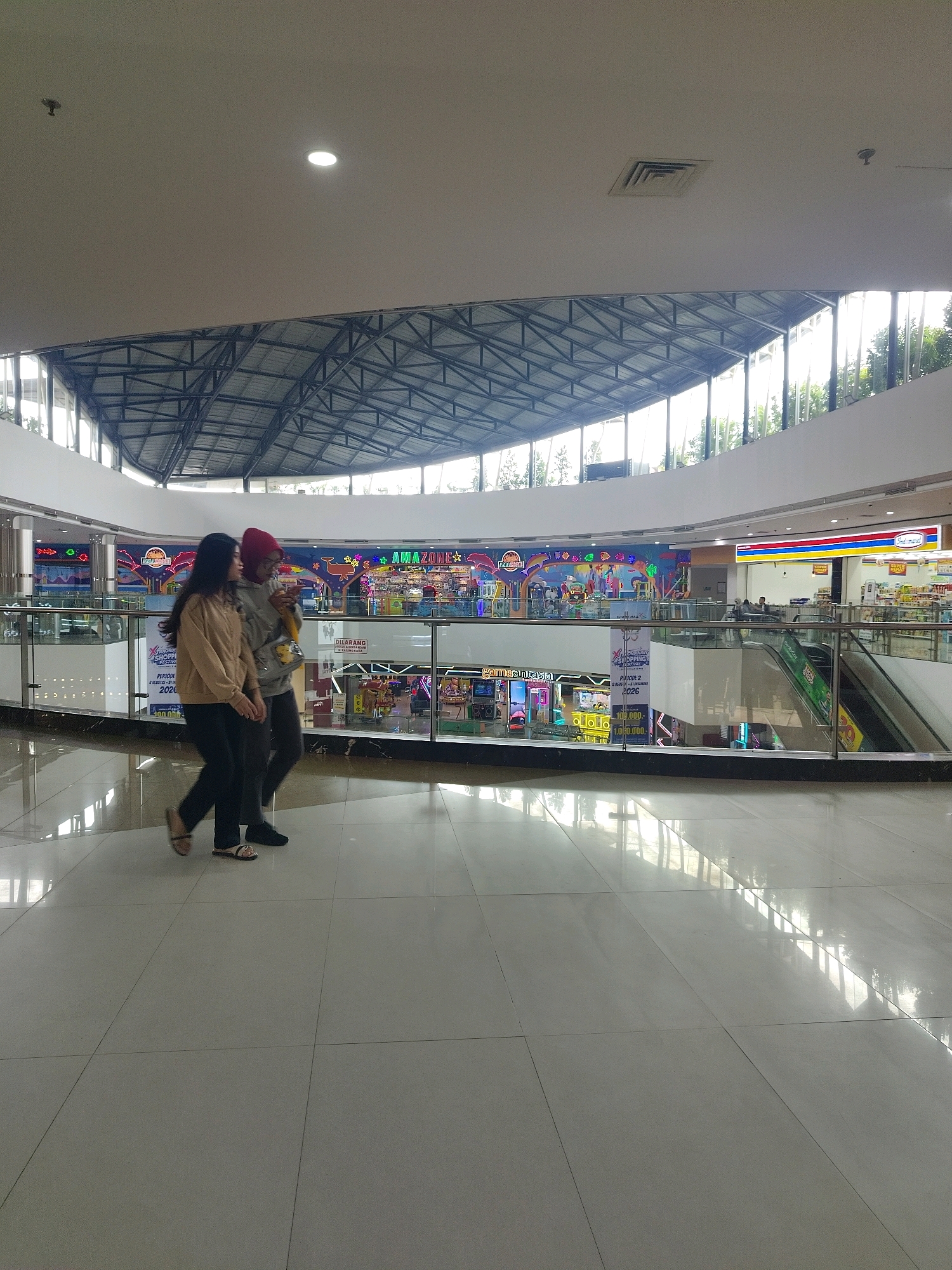 The image shows the interior of a modern shopping mall. Two young women are walking side-by-side in the foreground, on a polished tiled floor. The woman on the left has long dark hair and is wearing a tan sweatshirt and black pants. The woman on the right wears a red headscarf, a gray hoodie, and dark pants. They are walking towards the right side of the frame. In the background, on an upper level, there are various storefronts with bright signage, including what appears to be an arcade with colorful lights and a convenience store labeled "Indomaret". The mall features a multi-level design with glass railings and escalators visible. The architecture includes a prominent steel roof structure with translucent panels allowing natural light to enter. The overall environment is clean and well-lit. This scene is consistent with the interior of the Royal Plaza Mojokerto, a shopping mall located in the city of Mojokerto, Indonesia.