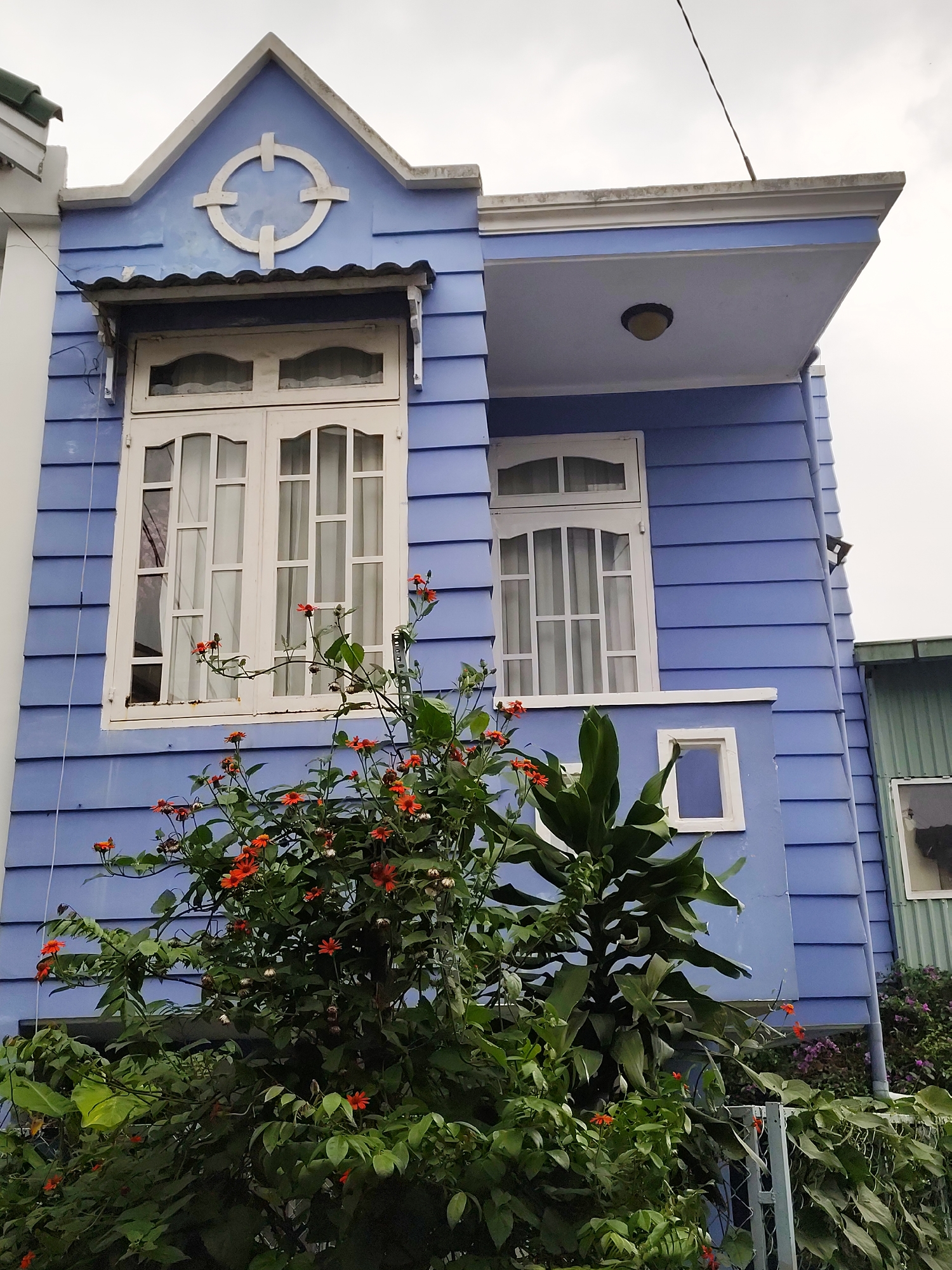 A two-story blue house with white trim is partially obscured by overgrown bushes with orange flowers. The house has horizontal siding and a gabled roof with a circular decoration. The lower window is large and divided into panes, while the upper window is smaller. The second story overhangs the first, supported by a small balcony. A small white square window is situated below the second window. The sky is overcast. The location is likely Da Lat, Vietnam, due to the architectural style and flora commonly found there.