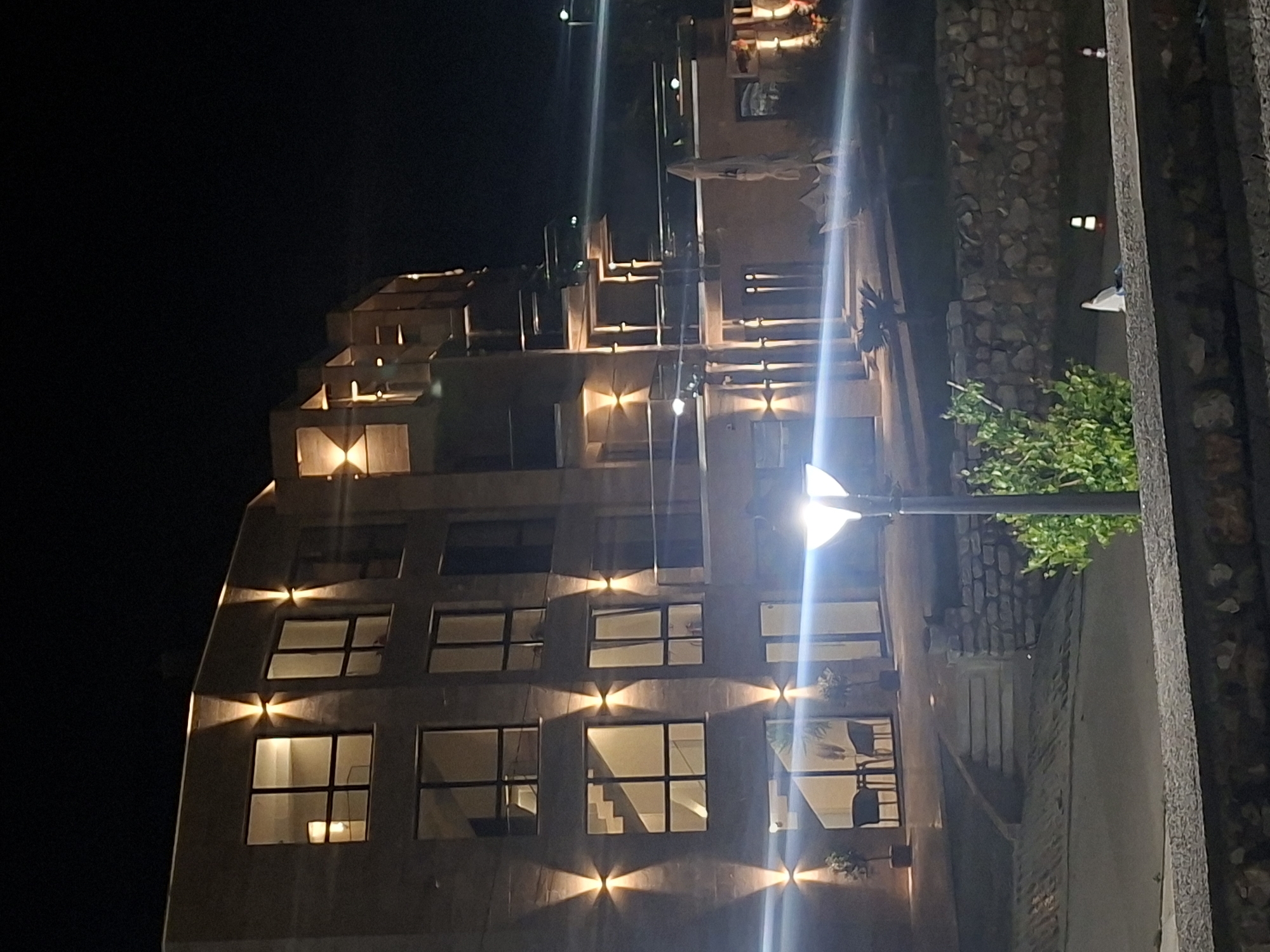 The image shows a modern, multi-story building at night in Tyulenovo, Bulgaria. The building has a facade of textured stone or brick and is illuminated by strategically placed uplights that highlight architectural features and windows. Several windows on the upper floors are lit from within, suggesting occupancy. On the ground floor, large glass doors lead into what appears to be an interior space with furniture visible, including chairs and tables. Outside, there are steps leading up to the building entrance, a stone wall, and landscaping with small trees. A modern lamp post with a bright light is positioned in the foreground, casting a significant glare across the scene. In the background, other lights are visible, and the dark sky suggests the late hour. The overall impression is one of a well-lit, contemporary establishment, possibly a hotel or restaurant, in a coastal or rural setting.