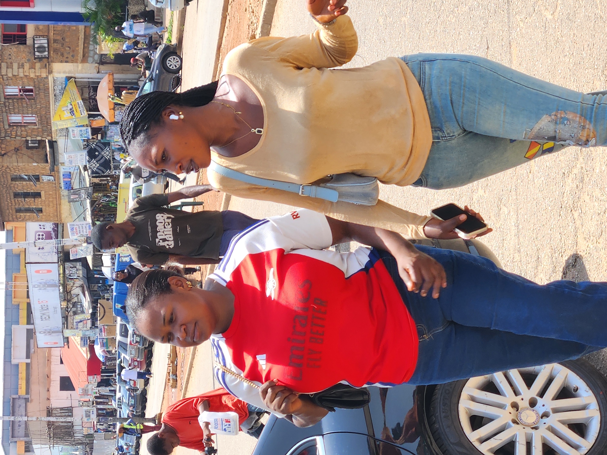The image depicts an outdoor scene in a bustling marketplace, likely in Jos, Nigeria. Two women are prominently featured, with their bodies angled towards the camera. The woman in the foreground, wearing a red and white shirt with "Emirates FLY BETTER" printed on it and blue jeans, looks downwards. Behind her, a woman in a light yellow long-sleeved shirt and blue jeans is looking to her right and holding a smartphone. She has an earring in her left ear and a necklace.
The background reveals a vibrant street scene with various elements of commerce. Buildings with brick and painted facades line the street, adorned with signs and posters, some advertising mobile phones like "Redmi Note 11". Several cars are parked along the road, and people are seen walking and interacting, suggesting a busy day. The ground is paved with gravel and dirt, typical of many urban streets in Nigeria. The lighting suggests it is daytime, with clear visibility. The overall atmosphere appears to be one of everyday activity and commerce in a lively urban environment.