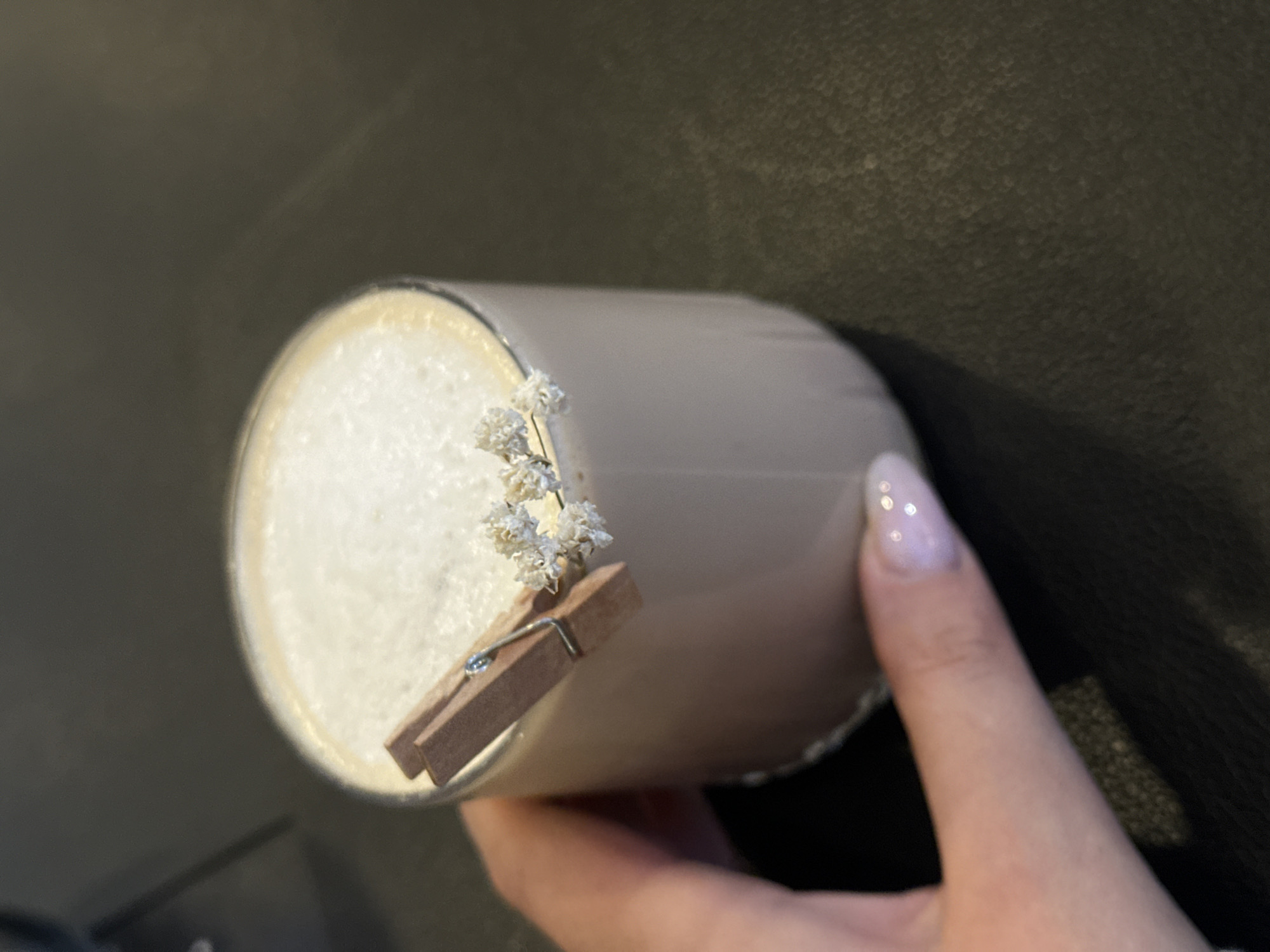 A hand holds a cylindrical glass filled with a white, foamy beverage, possibly a latte or cappuccino. The glass is adorned with a miniature wooden clothespin attached to a small sprig of dried white flowers. The hand features a manicured nail with a pale polish. The background is a textured, dark surface. There is no indication of the city of Šušanj, Montenegro, in the image.