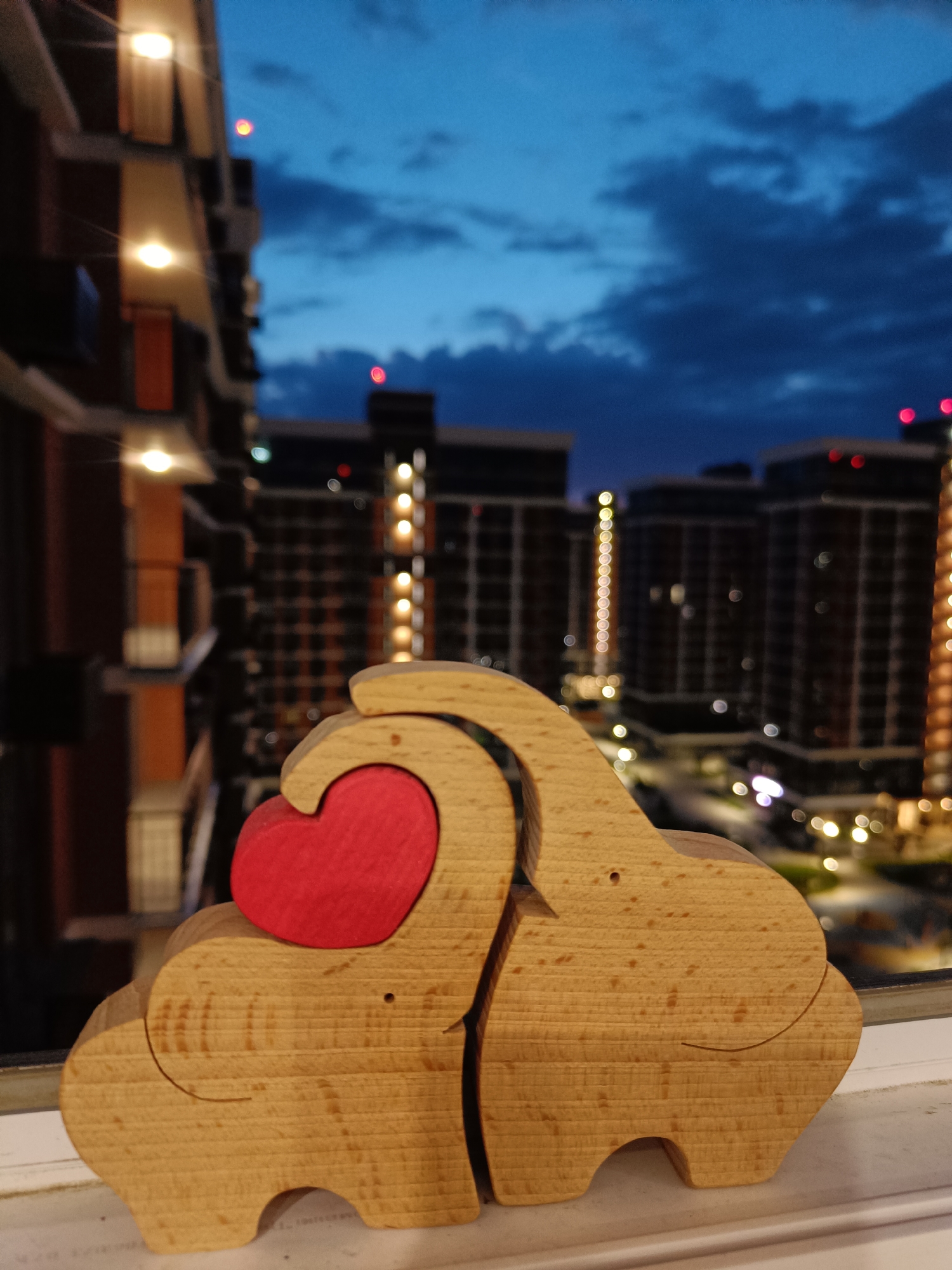 The image shows a wooden carving of two elephants with a red heart between them, placed on a windowsill. In the background, there are blurred images of apartment buildings with lights on, suggesting a city at dusk or night. The sky is dark blue with some clouds. There are no people visible in the image. The scene is likely taken from an apartment building, possibly in a residential area. The presence of apartment buildings with visible lights and modern architecture suggests an urban environment.