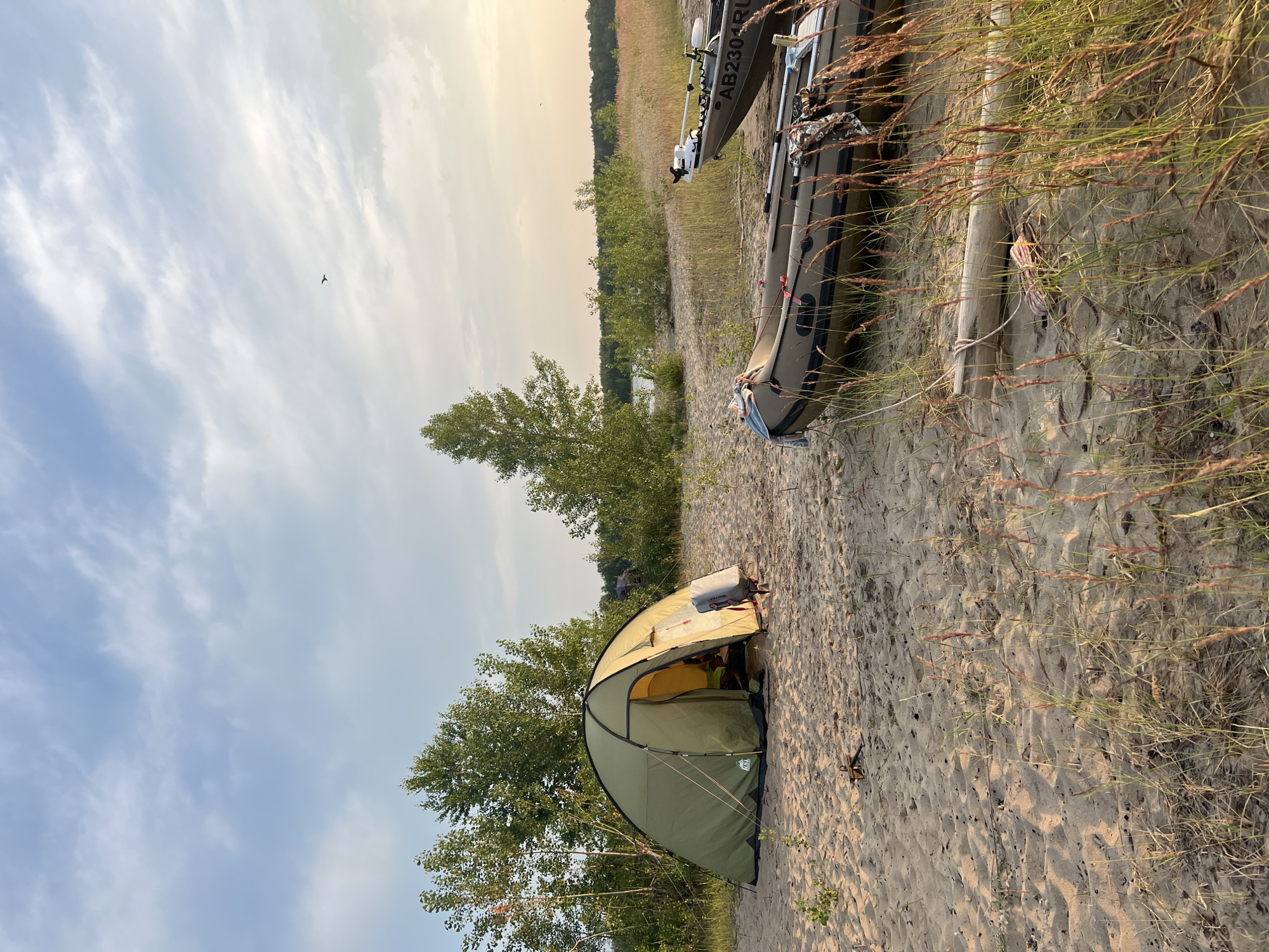 This is a photograph of a campsite on a sandy beach, likely located in Borovoe, Russia. The scene is illuminated by the soft light of late afternoon or early evening.
In the foreground, the sandy ground is covered with sparse, dry grass. Two inflatable boats are resting on the beach. The boat closer to the viewer is a dark olive green, and the one further back is a lighter gray with a visible registration number: "AB2304RU". Both boats appear to be equipped with electric trolling motors.
Dominating the left side of the image is a large, olive-green camping tent, partially open to reveal a yellow interior. A grey backpack is attached to the outside of the tent. Behind the tent, a line of trees forms a backdrop, with a body of water visible in the distance through gaps in the foliage. The sky above is a pale blue with wispy white clouds, and a small bird is visible in flight. The overall impression is one of a peaceful, remote camping and boating location.