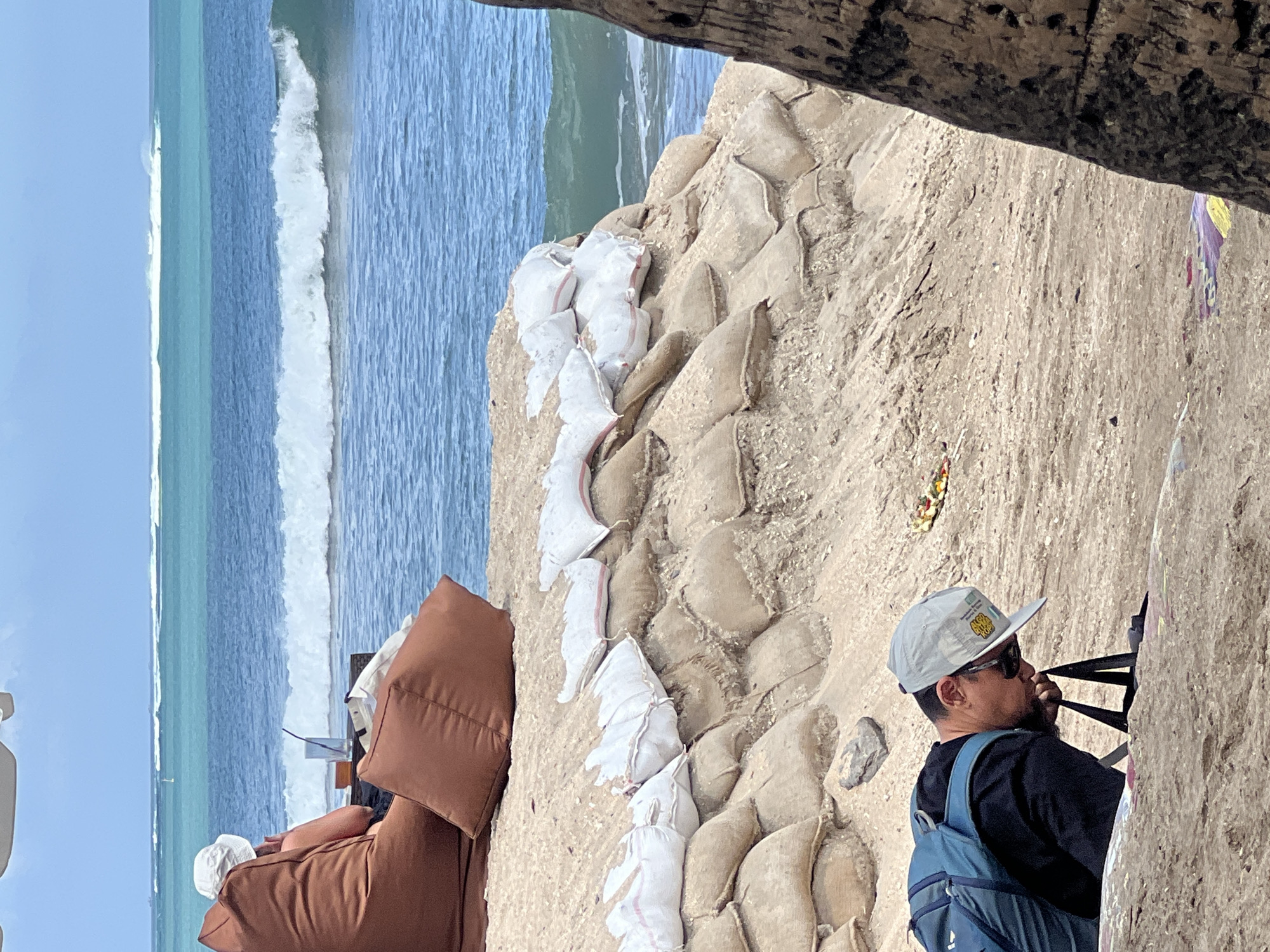 In Kuta, Indonesia, a man wearing a blue backpack, black shirt, sunglasses, and a white baseball cap is leaning forward on a sandy beach, appearing to be looking at something on the ground. Behind him, a fortified sandbag barrier extends along the coastline, topped with white sandbags marked with red stripes. In the background, the turquoise ocean meets the sky with visible waves. Further in the distance, two brown beanbag chairs are set up on the sand, with one person sitting in one of them, facing the ocean. The scene suggests a beach area prepared for coastal protection.
