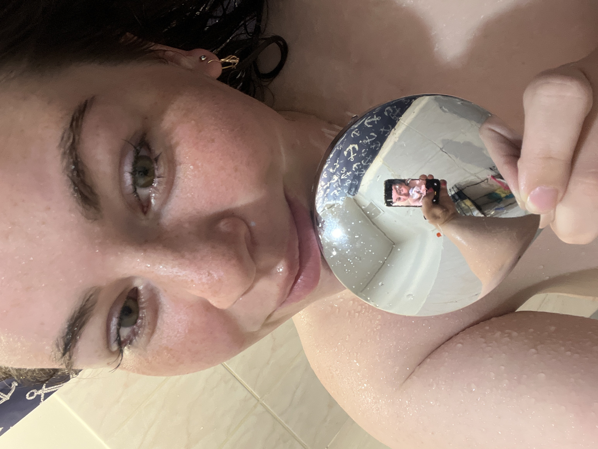 The image shows a woman's face and part of her upper body, wet with water droplets, likely from a shower. She is holding a round mirror that reflects a bathroom scene, showing a person holding a phone and taking a picture. The bathroom has light-colored tiles and a shower curtain with a nautical anchor pattern. The location, Chaikovsky, Russia, is not discernible from the image.