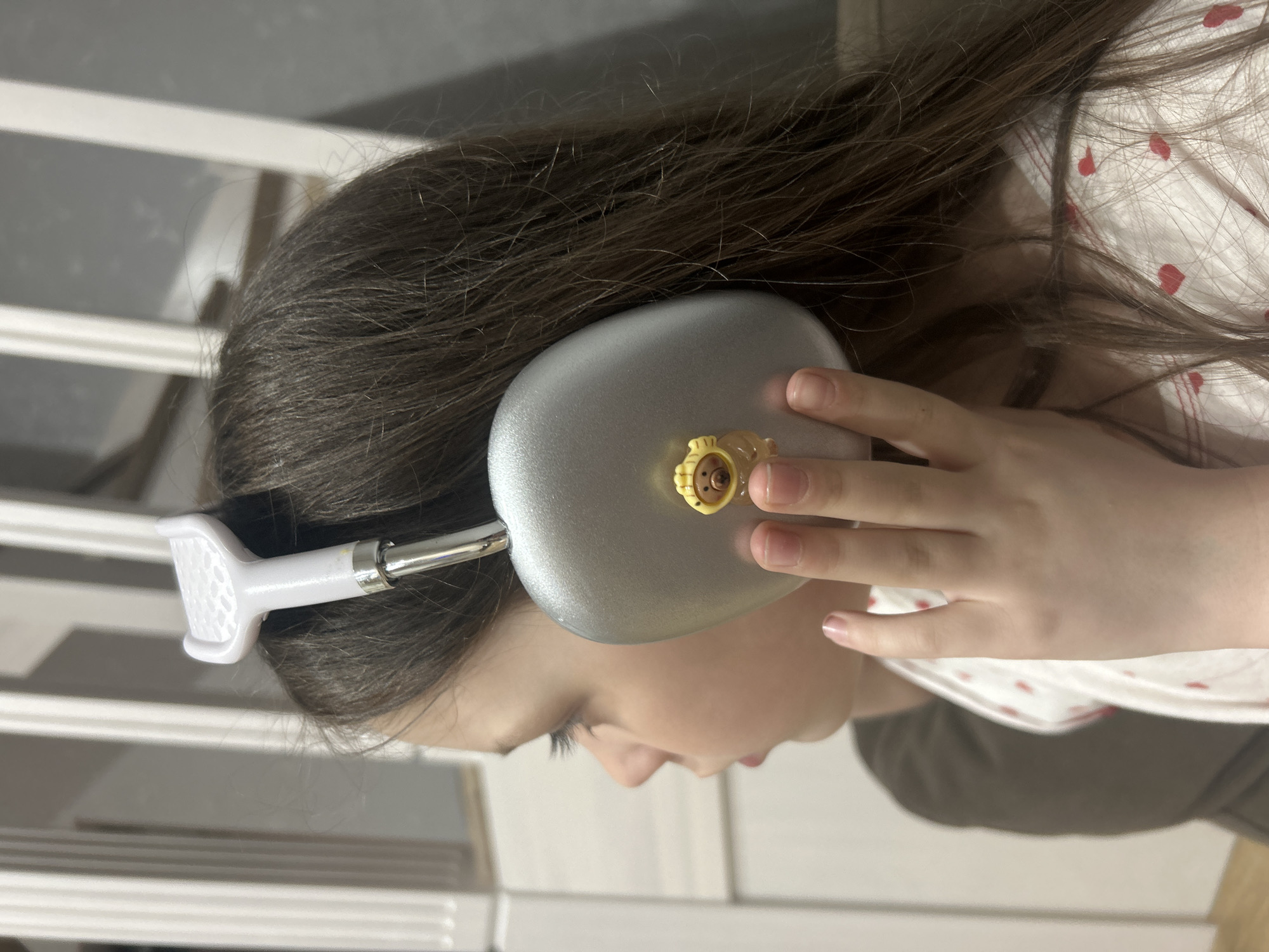 This close-up portrait features a young girl from a side profile, wearing silver over-ear headphones, in an indoor setting. Her long, dark brown hair cascades down, and her right hand gently rests on the headphone cup. A small, yellow, lion-like charm is attached to the exterior of the right headphone cup. She is dressed in a white top patterned with small red hearts, with a darker garment visible underneath. The headphones have a white headband with a textured section and a metallic extension connecting to the earcups. The background is softly blurred, revealing light-colored horizontal lines, possibly from furniture such as a crib or bed frame, or a wall panel, suggesting a domestic interior. The girl's expression is neutral, indicating focus or relaxation while listening. There is no visible text in the image. The lighting is soft and even, not indicating a specific time of day.