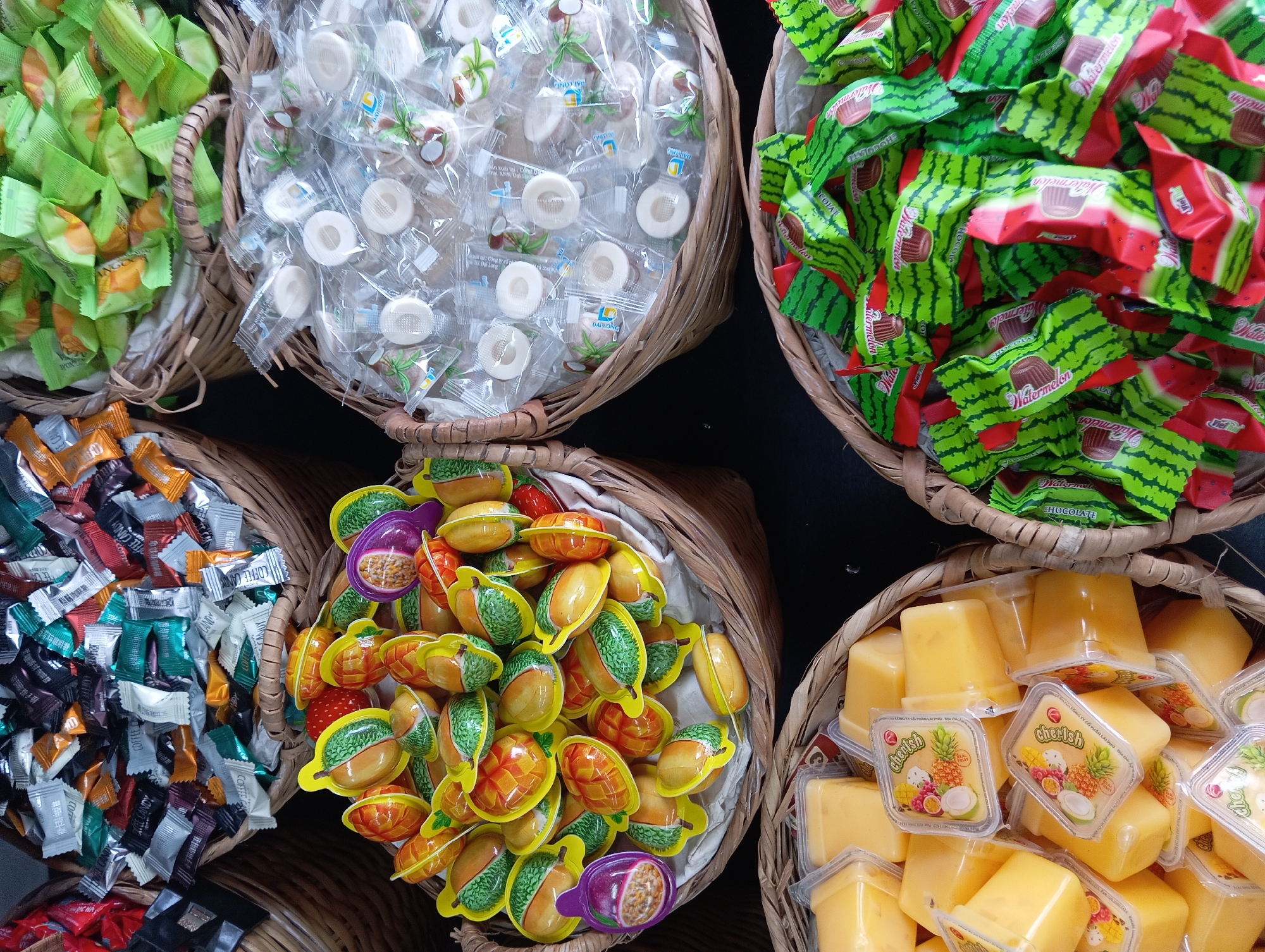 A close-up, overhead shot displays an assortment of candies and desserts arranged in wicker baskets. In the top left, a basket overflows with individually wrapped candies in various colors, including red, orange, silver, black, and green, some featuring text like "COFFEE CANDY". To the right of this, another basket is filled with green-wrapped candies, some displaying orange or yellow fruit imagery.
Below the first two baskets, a larger basket holds candies shaped like fruits, predominantly yellow with green accents, resembling mangoes or durians, interspersed with a few purple passion fruit-shaped candies. To the right of these fruit candies, a basket contains numerous small, clear-wrapped candies, many with white circular candies inside, some showing images of coconuts.
In the bottom left, a basket is filled with small, square, clear-plastic containers of a yellow, custard-like dessert, each topped with a lid featuring images of tropical fruits like pineapple, mango, and passion fruit, and the text "Checish". To the right of these desserts, a basket is packed with candies wrapped in bright green and red packaging, designed to resemble watermelon slices, with some wrappers showing text like "Watermelon" and "CHOCOLATE". The overall impression is of a vibrant, sweet display, likely in a market or shop. This scene could be located in Nha Trang, Vietnam, a city known for its street markets and sweet treats.