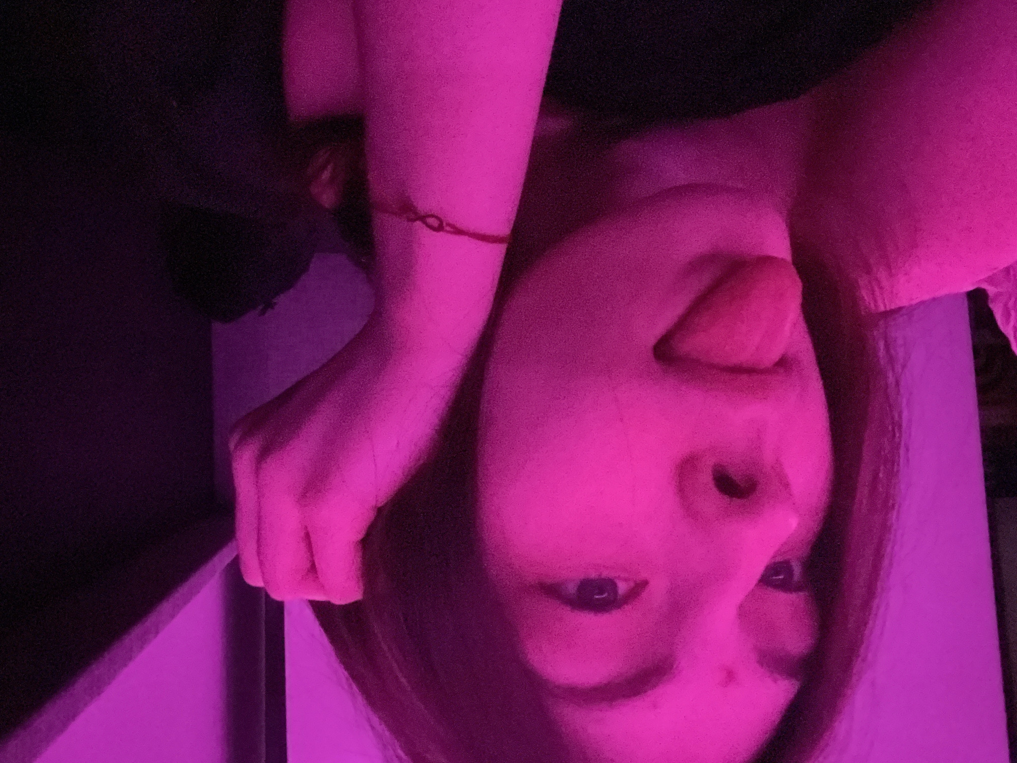 A close-up portrait of a person is depicted, rotated at an unusual angle, making the subject appear inverted. The scene is illuminated by strong pink and magenta artificial lighting, casting deep shadows and coloring the skin and surroundings in vibrant fuchsia tones. The person's face is central, with a playful expression, as they stick their tongue out towards the viewer. Their eyes, visible as dark pupils against the pink-tinted face, engage directly with the camera.
One arm is raised and bent, with a thin red string or bracelet visible on the wrist. The hand appears to be clenched or grasping something, though it's largely obscured by shadow and light. The background consists of dark areas, likely walls or furniture, interspersed with patches of pink light. The overall setting appears to be indoors, likely at night or in a room with controlled lighting. The image has a grainy texture, suggesting low-light photography. No text is visible.