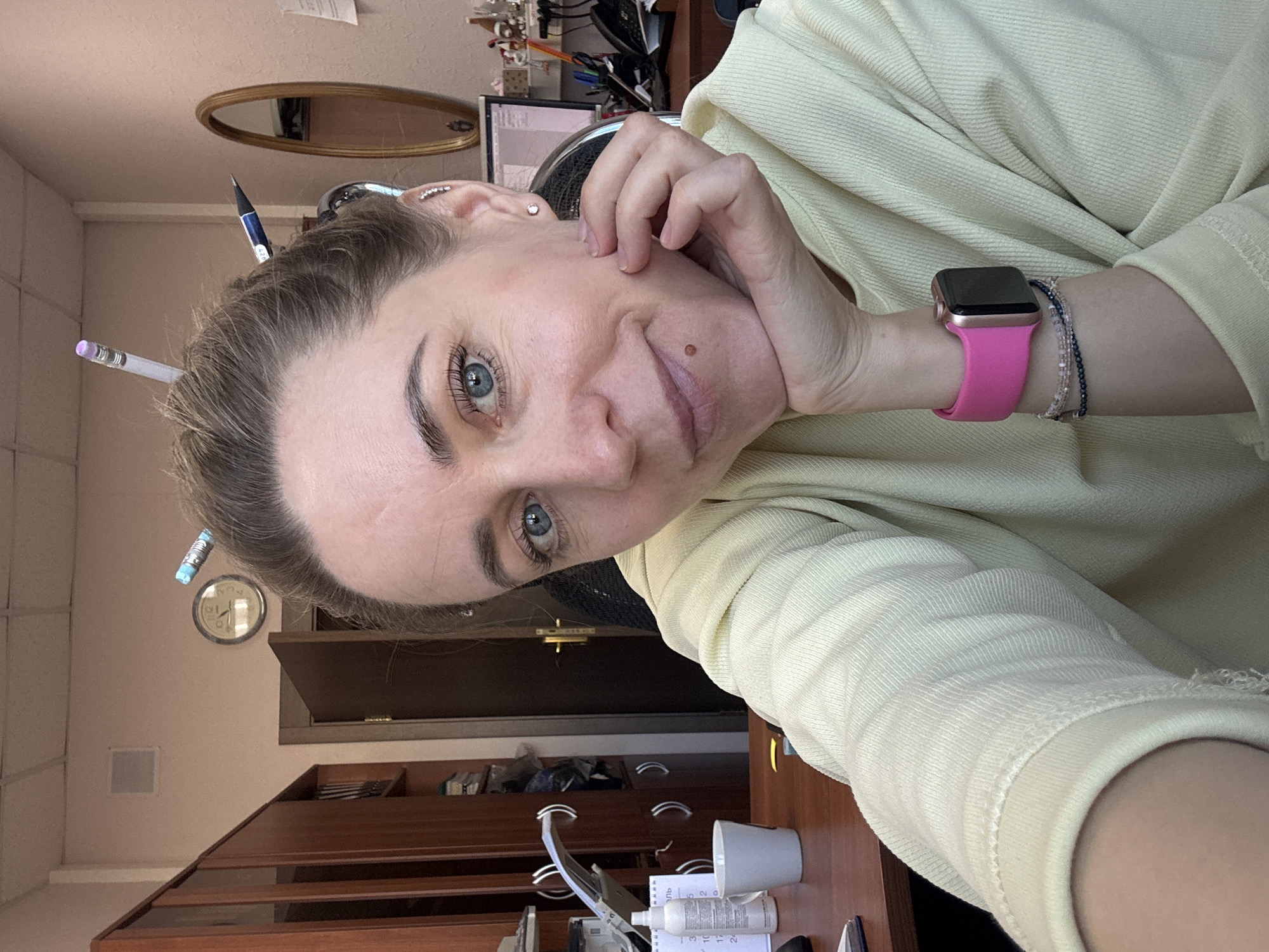 A selfie shows a woman with light skin, blue eyes, and light brown hair with pencils sticking out of it. She's wearing a pale yellow sweatshirt and a pink Apple Watch. Her face is turned slightly towards the camera, with her hand resting on her cheek. The background includes a desk with a cup, bottles, and computer equipment. Also visible are a brown wooden cabinet, a round clock on the wall, and a framed oval mirror. The scene may be in an office setting located in the city of Chaikovsky, Russia.