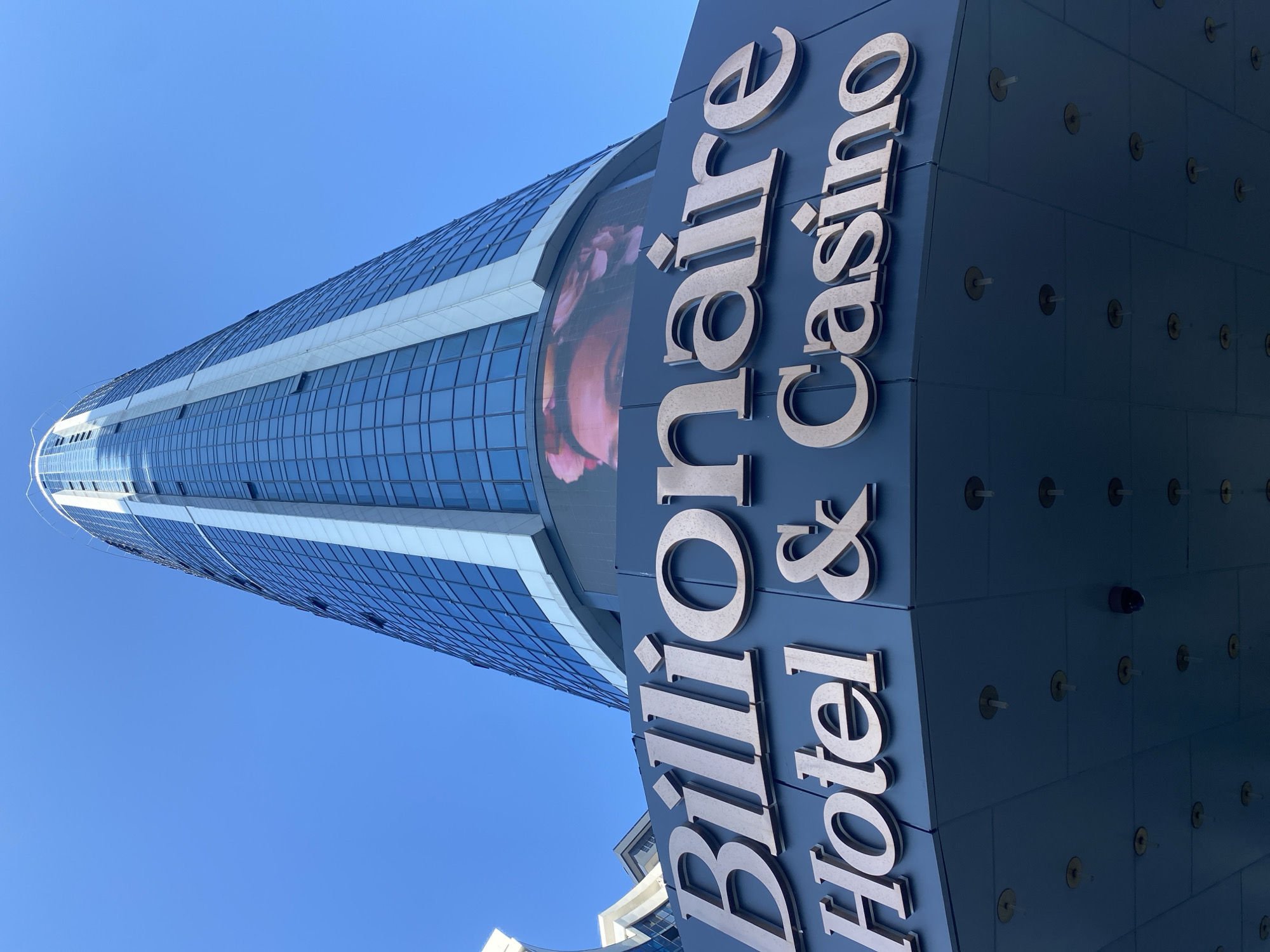 The image captures the "Billionaire Hotel & Casino" building in Batumi, Georgia, as viewed from a low angle looking upwards against a clear blue sky. The building is a modern skyscraper with a facade primarily composed of blue glass, reflecting the sky. A prominent sign with the words "Billionaire Hotel & Casino" in large, metallic 3D lettering is displayed on the dark blue lower section of the building, which also features a large digital screen displaying a close-up of a person's face. The underside of this section of the building has a grid of circular fixtures and at least one visible security camera.