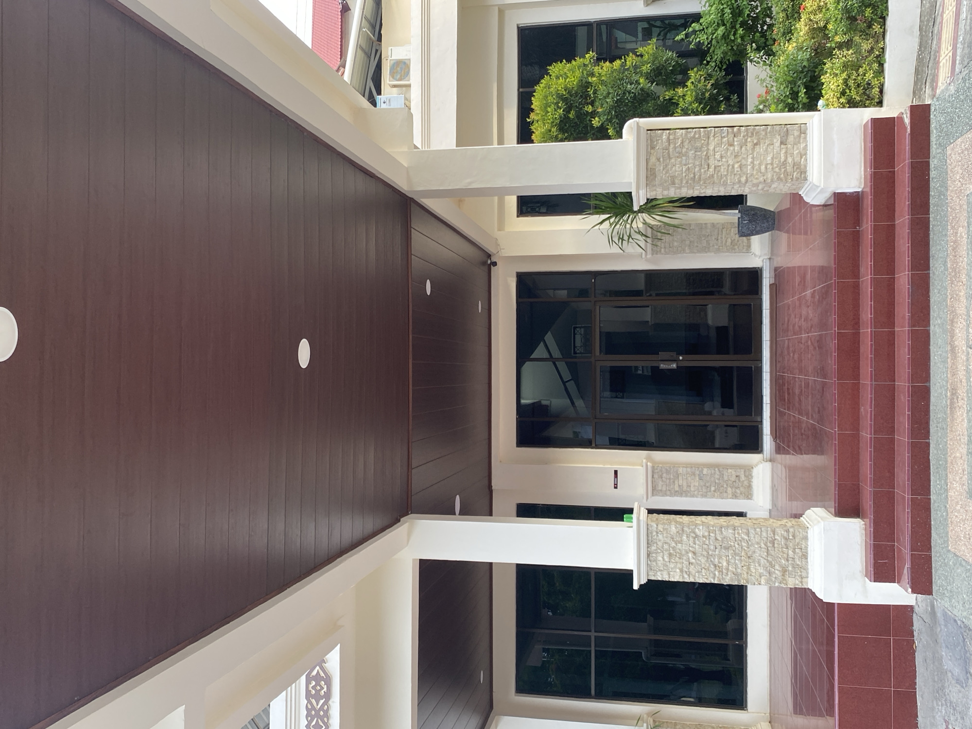 The image shows the entrance to a building in Banda Aceh, Indonesia. The entrance features a covered walkway with a dark brown, wood-paneled ceiling and white columns. The columns are square and have a textured stone finish on the lower half. There are large glass doors in the center of the entrance, flanked by large windows on either side. A set of red tiled stairs leads up to the entrance. To the right of the entrance, there is a lush green bush and a potted plant. The building appears to be modern and well-maintained. The overall impression is one of an inviting and professional establishment.