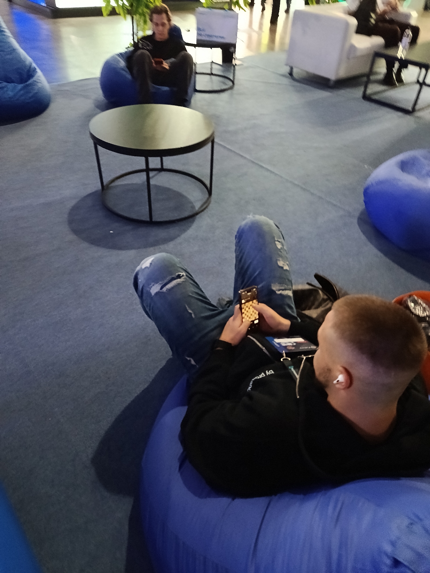 The image shows a person reclining on a blue beanbag chair in what appears to be an indoor lounge or waiting area. The person is wearing ripped blue jeans and a black hoodie, and they are holding and looking at a smartphone. Another person is sitting in a beanbag chair in the background, also looking at a phone. In the background, there's a small round table and a white armchair. The floor is covered with blue carpeting. Based on the information available, it is not possible to conclusively determine that the location is in Khimki, Russia.