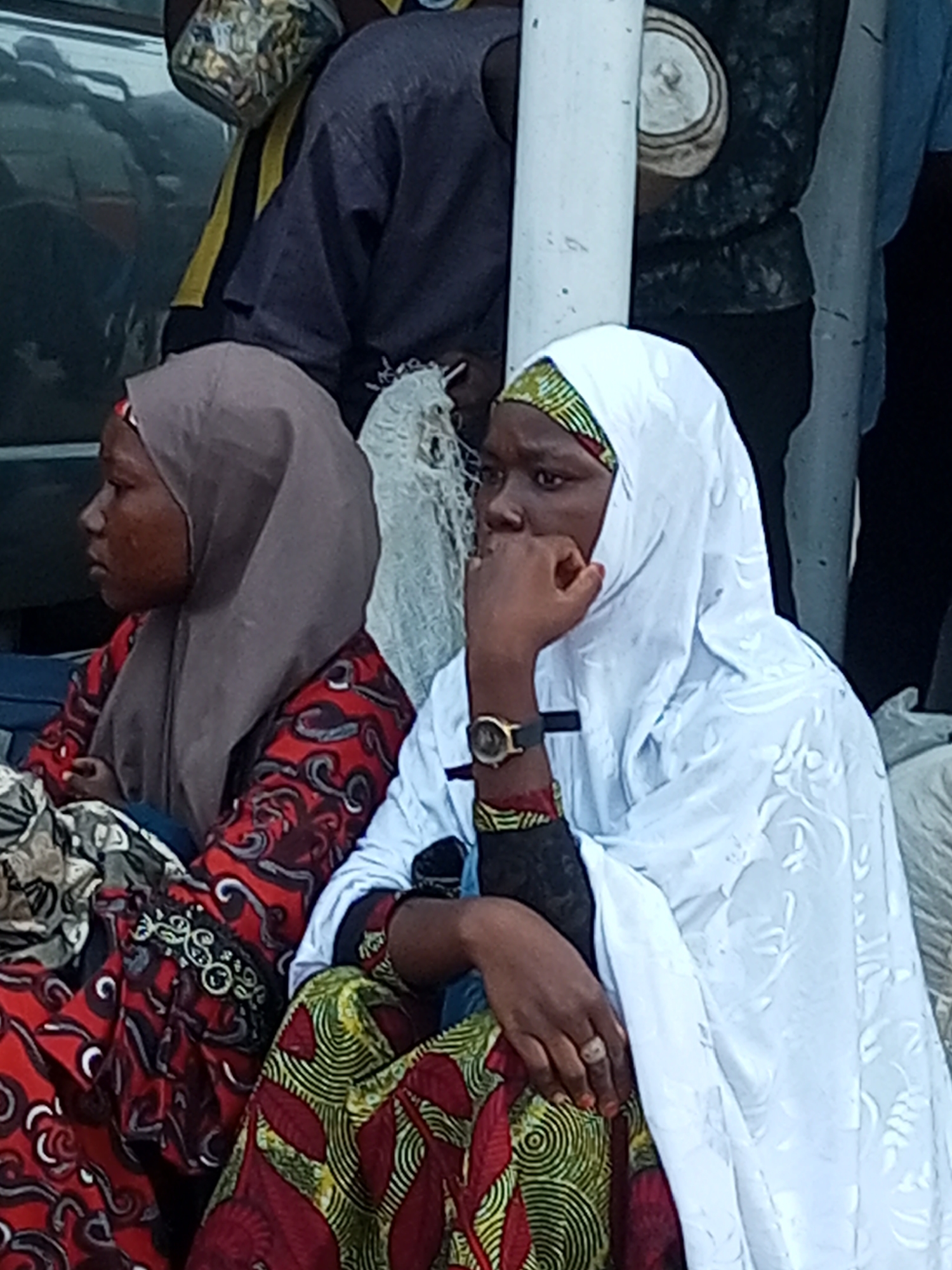 A medium shot shows two young women in vibrant traditional African attire seated next to each other. The woman on the left, wearing a brown hijab and a red patterned dress, is seen from the side, her head tilted down, appearing pensive. The woman on the right is facing forward, wearing a white hijab and a dress with a green and yellow circular pattern. Her right hand is raised to her chin, and she wears a wristwatch with a black strap. They are seated outdoors in what appears to be a public space within the city of Gombe, Nigeria. The background is blurred, suggesting a busy environment, with hints of other people and structures. The lighting is natural, indicating it's daytime.