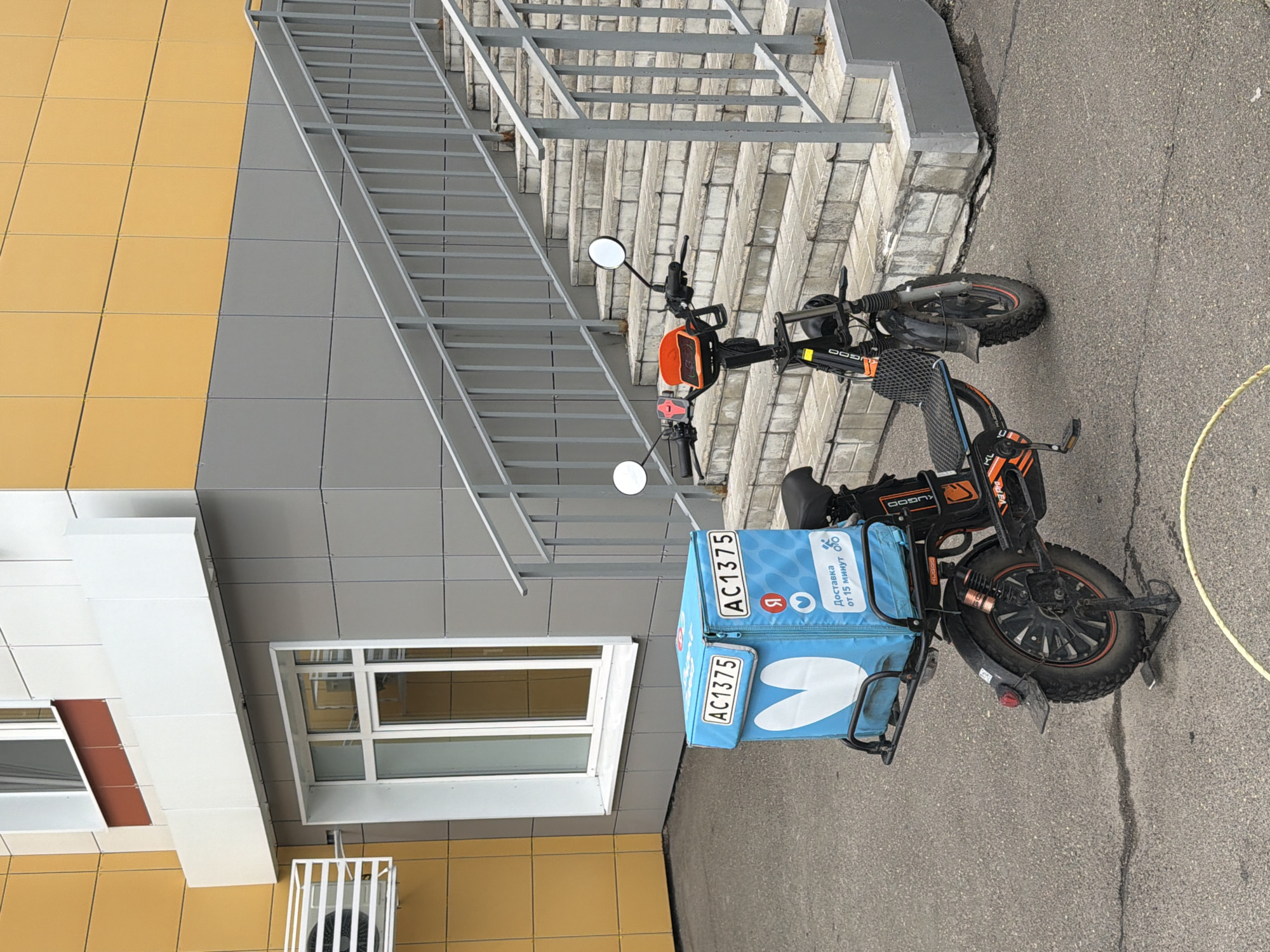 This image shows an electric scooter parked on an asphalt surface next to a modern building with yellow and gray tiled exterior walls. A metal staircase with railings runs alongside the building. The scooter has a large blue insulated delivery bag attached to its rear, with white text and logos on it, including "AC1375", a red "Я" logo, and "Доставка от 15 минут". The scooter itself is black and orange and appears to be a delivery vehicle. The scene is outdoors, with no people visible. The lighting suggests it is daytime. The building has several windows. The overall impression is one of an urban or commercial setting where delivery services are active.