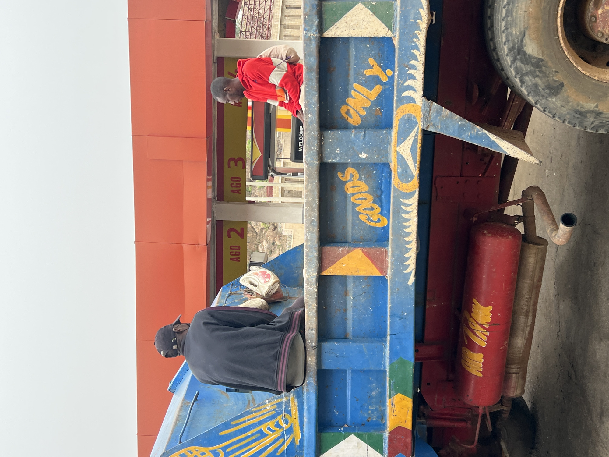 The image shows a brightly painted truck in what appears to be Jos, Nigeria, based on possible AGO fuel station signs in the background. Two men are present. One in a red and white tracksuit leans over the truck's side facing the station. The other man, wearing a dark hat and dark clothing, is sitting on the truck bed, next to a white object. The truck bed is painted blue with geometric patterns and the words "GOODS ONLY." A spare tire and red air tank are visible on the truck. The sky is overcast.
