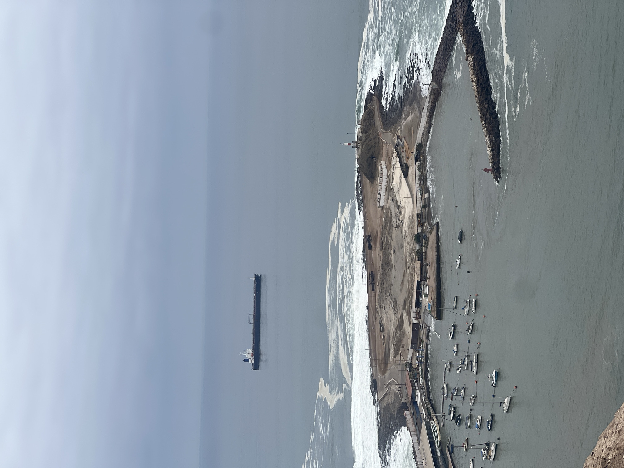 This image provides an elevated, wide-angle view of a coastal area identified as Arica, Chile, under an overcast sky. The upper half of the image features a uniform, light grey sky meeting a calm, grey-blue ocean at the horizon.
In the mid-distance on the open ocean, a large cargo vessel, characterized by a dark hull and a white and blue superstructure, is visible. Closer to the foreground, a prominent rocky peninsula extends into the sea. On the summit of this peninsula stands a red and white striped lighthouse. The outer perimeter of the peninsula and the adjacent coastline are subjected to significant white foamy waves breaking against the rocky shore. Several low-lying structures or foundations and a dirt road or path are observable on the barren terrain of the peninsula, with a few small human figures discernible walking on these paths.
To the left of the peninsula, a protected harbor is formed by a curved, segmented rock breakwater. A small red beacon or navigational light is situated at the seaward end of this breakwater, marking the harbor entrance. Inside the tranquil waters of the harbor, numerous recreational boats, including both sailboats and motorboats, are moored. Along the inner shore of the harbor, several buildings and structures are visible, consistent with maritime facilities. The immediate foreground, at the bottom left, shows the edge of a reddish-brown rocky cliff, indicating the elevated position of the viewpoint.