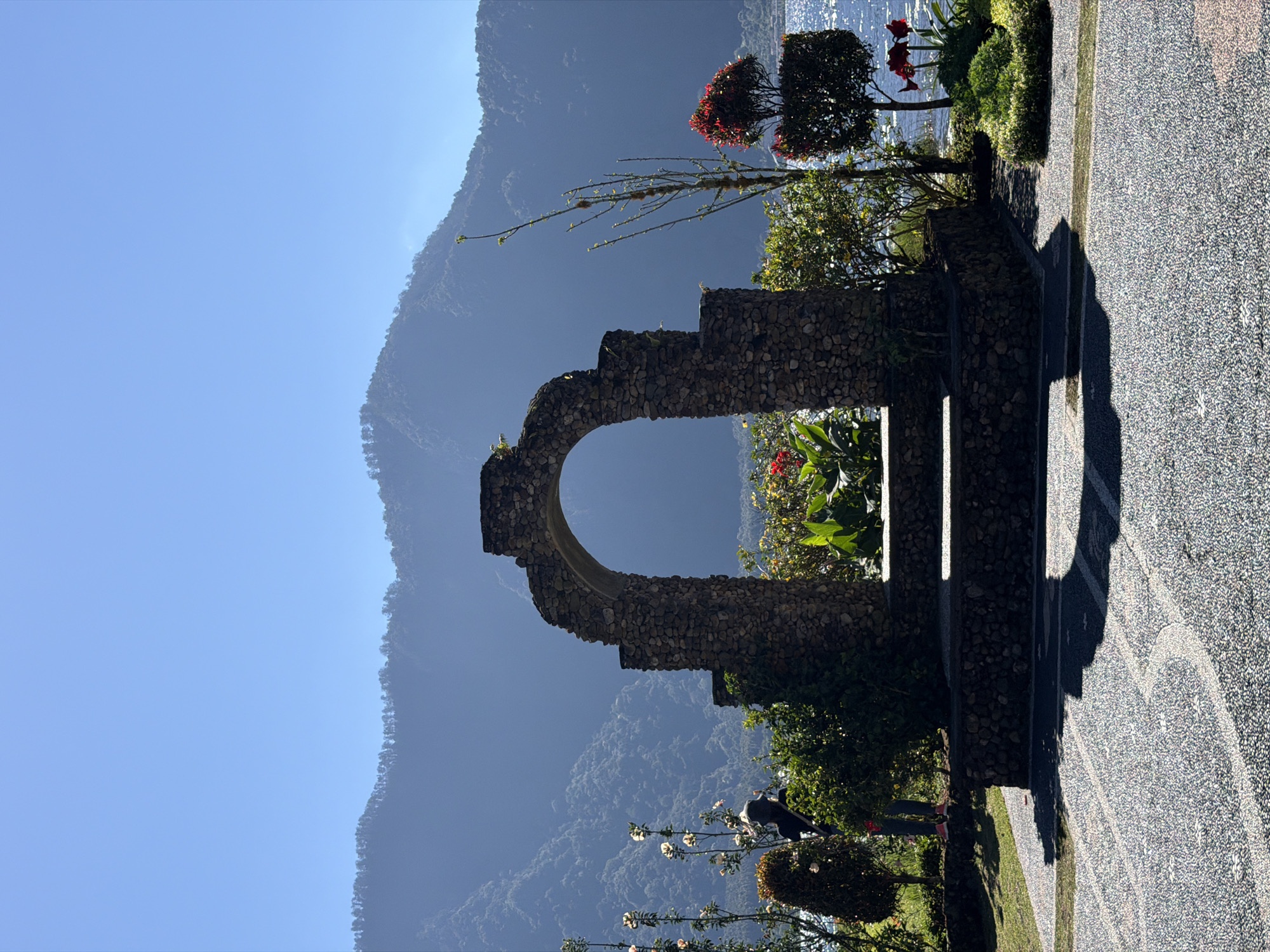 In Baturiti, Indonesia, a stone archway with a rustic, natural stone finish stands prominently in the foreground. It frames a lush garden area with vibrant green plants and red flowers, leading to a serene body of water, likely a lake or ocean, visible in the background. The water is calm and reflects the clear blue sky. Beyond the water, a majestic mountain range rises, its slopes covered in dense vegetation and softened by a gentle haze, creating a sense of depth and grandeur. A person, silhouetted against the bright sky and mountains, is partially visible on the left side of the archway, appearing to be looking towards the scenery. The foreground is paved with a textured, light-colored material, likely a pathway or courtyard, with shadows from the archway and surrounding greenery cast upon it. The overall scene conveys a tranquil and scenic atmosphere, blending natural beauty with architectural elements.