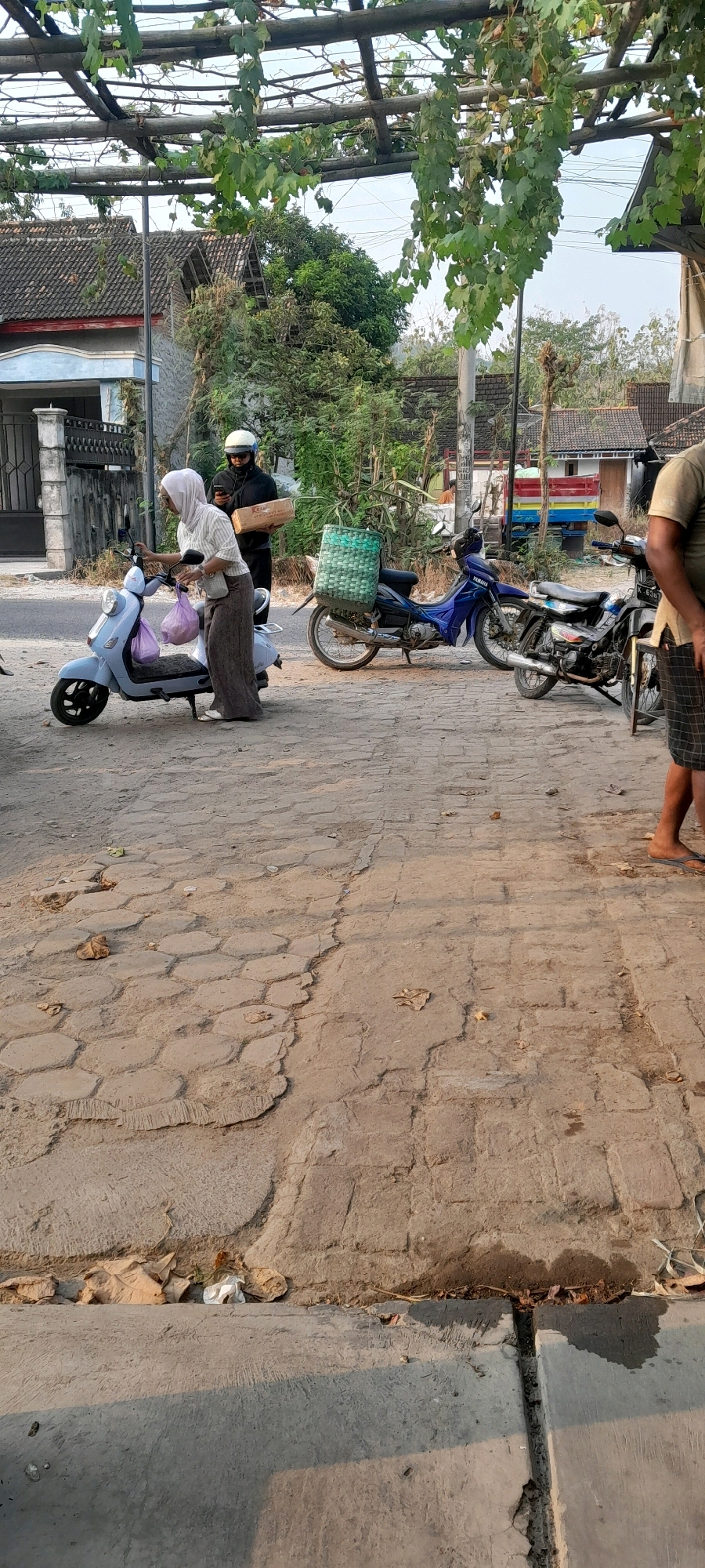 In Sambong, Indonesia, a woman in a headscarf and patterned trousers is reaching for a purple bag on the seat of a light blue scooter. She is standing on a textured, reddish-brown paved surface. Behind her, a person wearing a dark jacket and helmet holds a rectangular package. To their right, a blue motorcycle with a large green woven basket on its rear is parked. Further to the right, another motorcycle is partially visible. The background shows residential buildings with tiled roofs and lush green foliage. The scene appears to be outdoors, possibly on a street or in a yard, with the sunlight casting shadows.