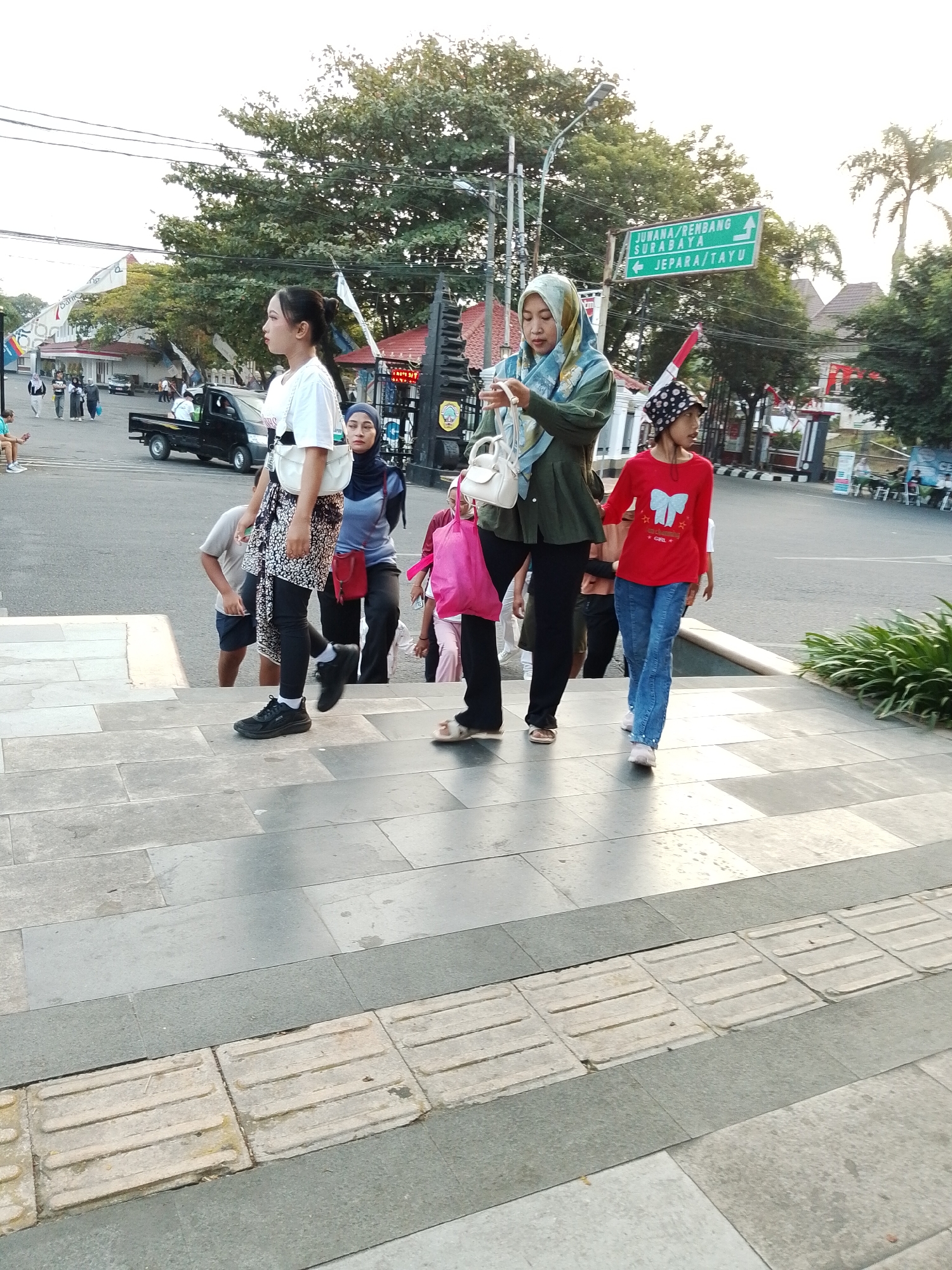 In the city of Kecamatan Pati, Indonesia, a group of people are walking up a set of outdoor stairs. A young woman in a white t-shirt and patterned skirt is walking up the stairs, looking to her left. Behind her, a woman in a green hijab and a white purse is looking at her phone. To the right, a young girl in a red shirt and blue jeans walks up the stairs. A pink bag is visible on the stairs, held by a person further behind. In the background, a street is visible with a truck and traffic signs. The environment appears to be an urban outdoor setting with trees and buildings.