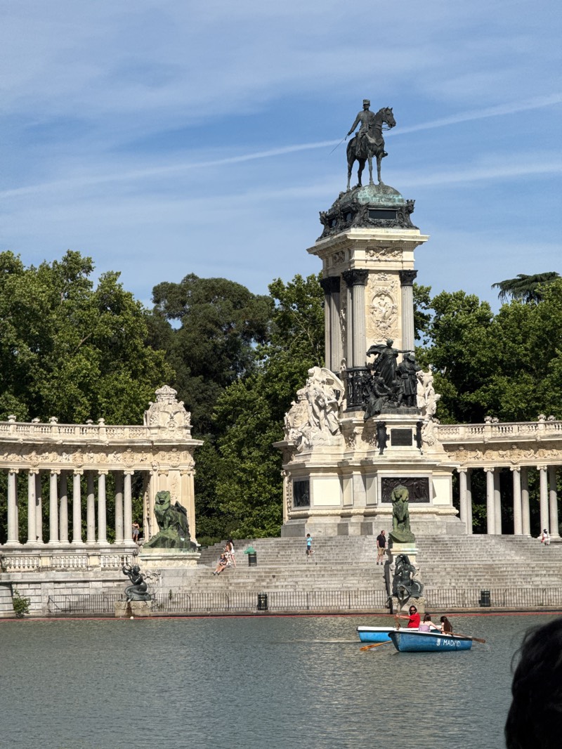The image presents a vibrant daytime scene at the Monument to Alfonso XII in El Retiro Park, Madrid, Spain. The grand monument dominates the center, featuring a large equestrian statue of King Alfonso XII atop an ornate, multi-tiered pedestal, decorated with sculptures, columns, and architectural details. This central structure is flanked by an expansive semicircular colonnade with classical pillars and further sculptures, leading down to a wide, terraced staircase.
In the foreground, a calm body of water, likely the park's lake, reflects the sunlight. Several small, blue rowboats are visible on the water, carrying people enjoying a leisurely paddle. One boat, towards the right, clearly displays the word "MADRID" and the number "3" on its side, with multiple occupants including one person rowing.
People are scattered across the monument's steps and terraces, some sitting and relaxing, others walking. Lush green trees frame the background, indicating a park setting. The sky is bright blue with a few faint, wispy clouds, suggesting clear, sunny weather, likely midday or early afternoon given the illumination. The overall scene depicts a peaceful and popular recreational area, combining historical architecture with natural beauty and human activity.