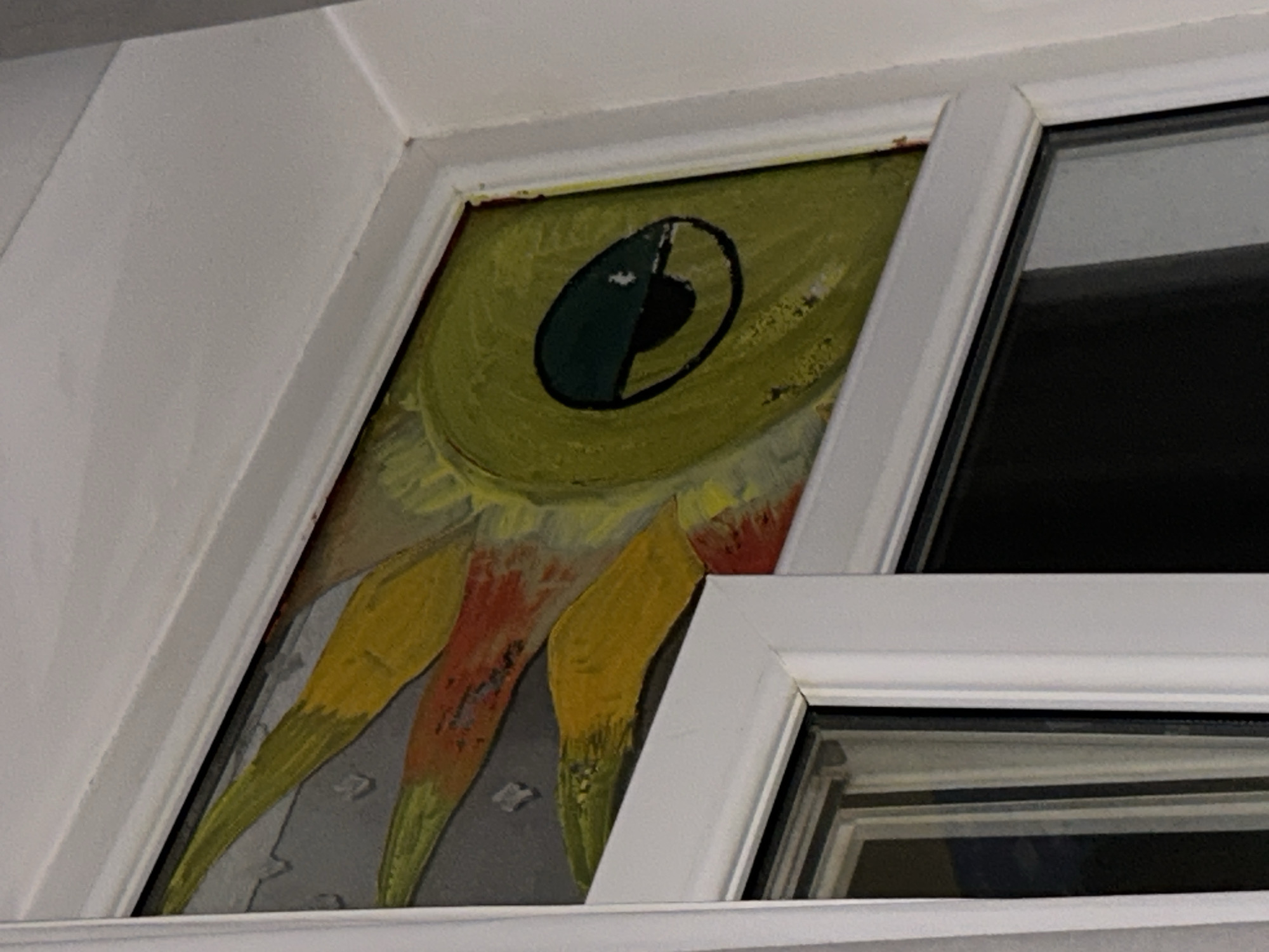 A close-up, eye-level, exterior shot captures a section of a white window frame. Within the upper portion of the frame, a painting or drawing is visible. The artwork depicts a stylized, cartoonish eye, predominantly yellow with a teal pupil and black outline. Surrounding the eye are pointed, sun-like rays in shades of yellow, orange, and red, painted against a gray, textured background that appears to represent a wall. The painting is slightly askew within the window opening. Below the painted section, the image shows the interior of a modern double-paned window with black frames. The lighting suggests it may be nighttime or indoors with artificial light. The location is described as Unknown, Unknown.